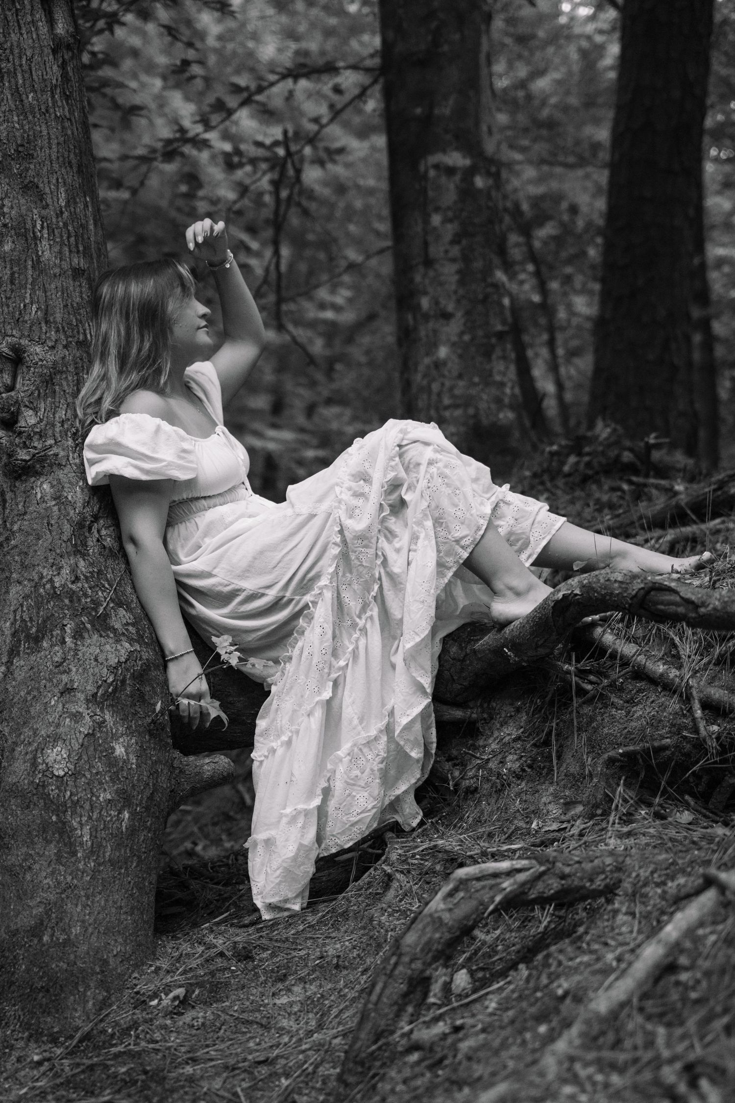 Black and white photo of a woman in white dress reclining against a tree in the woods.