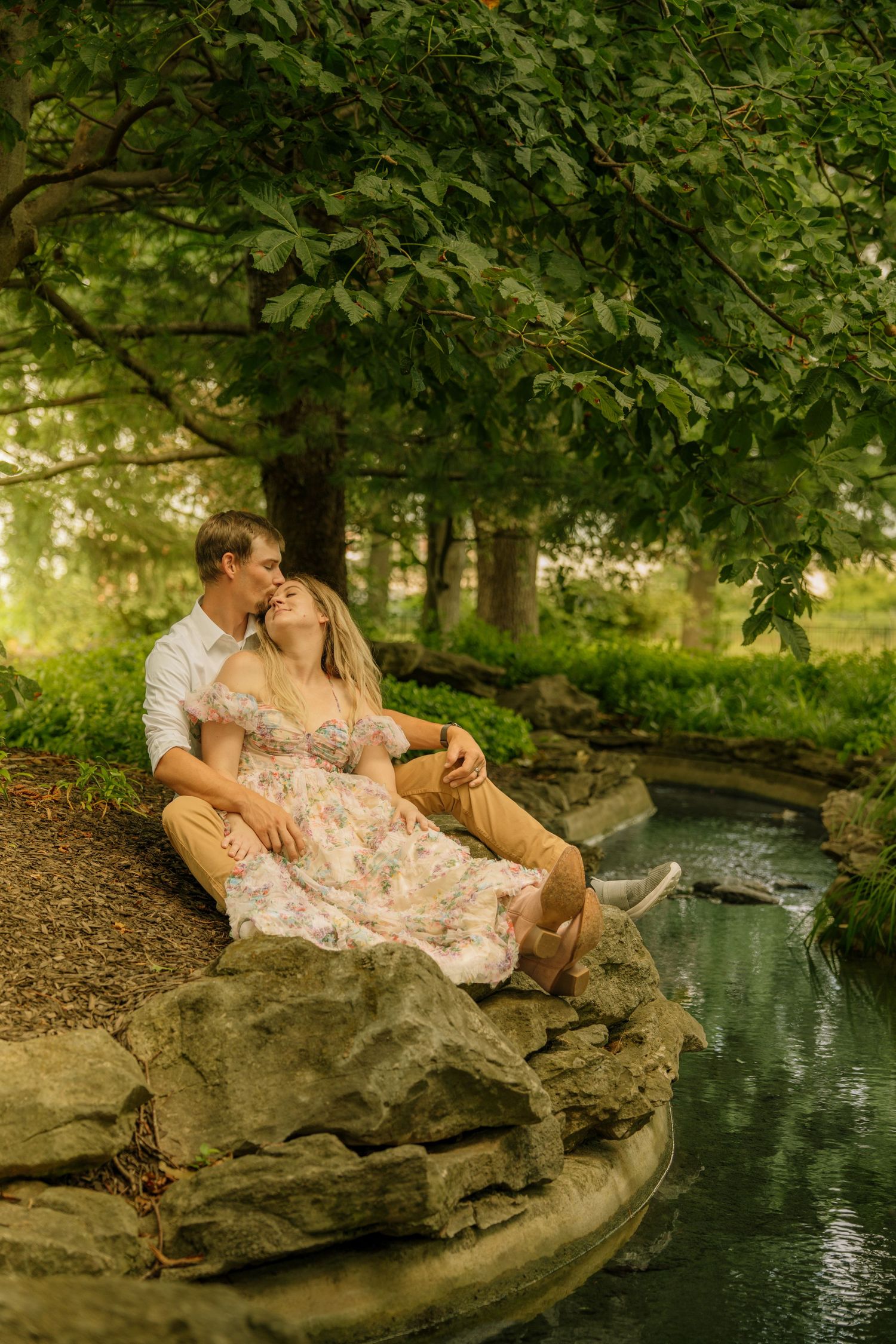 Couple sitting together on rocks by a stream under lush trees, sharing an intimate moment at golden hour.