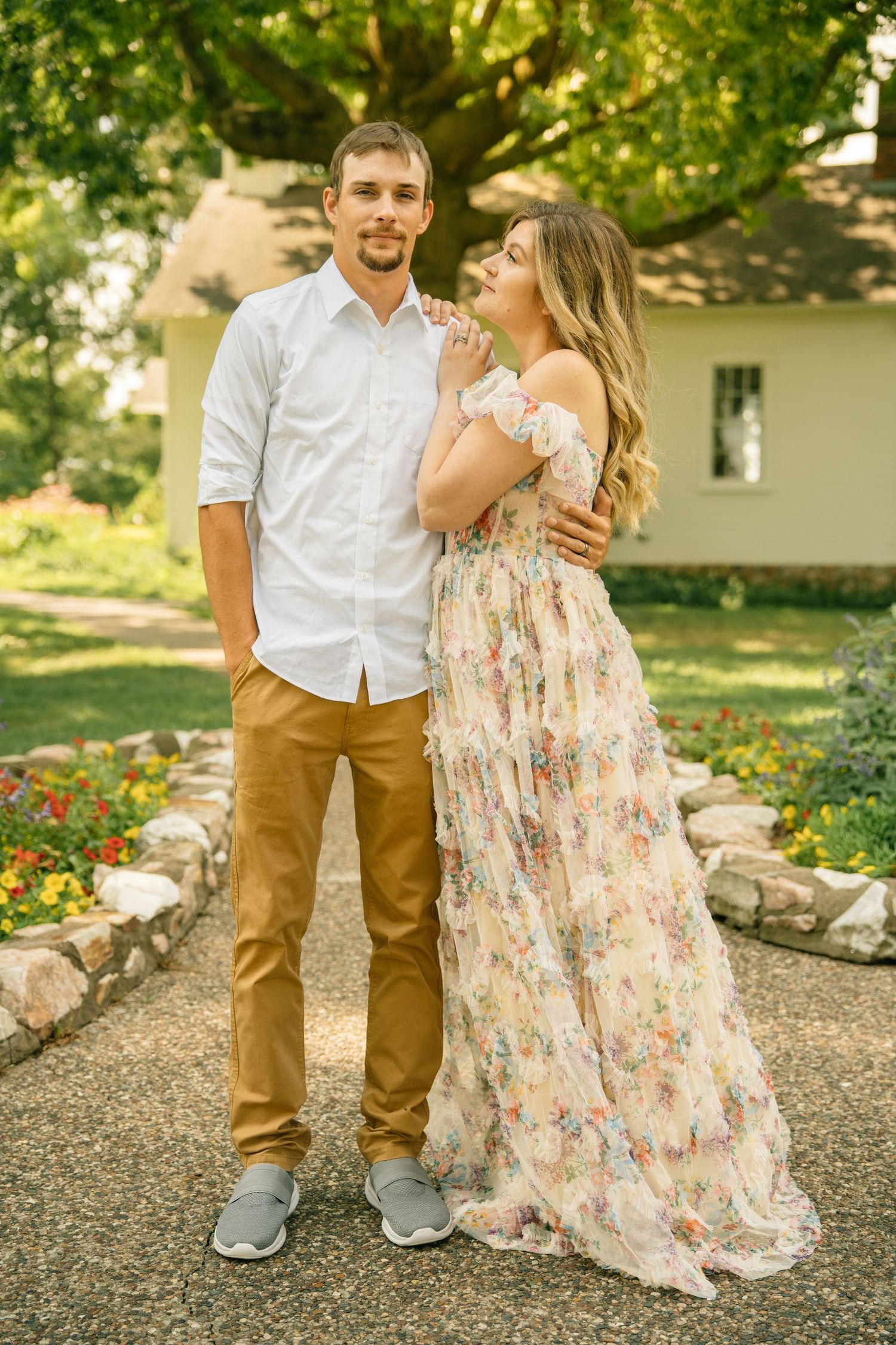 Couple posing in a garden, man in white shirt and tan pants, woman in floral maxi dress, with greenery and flowers behind.