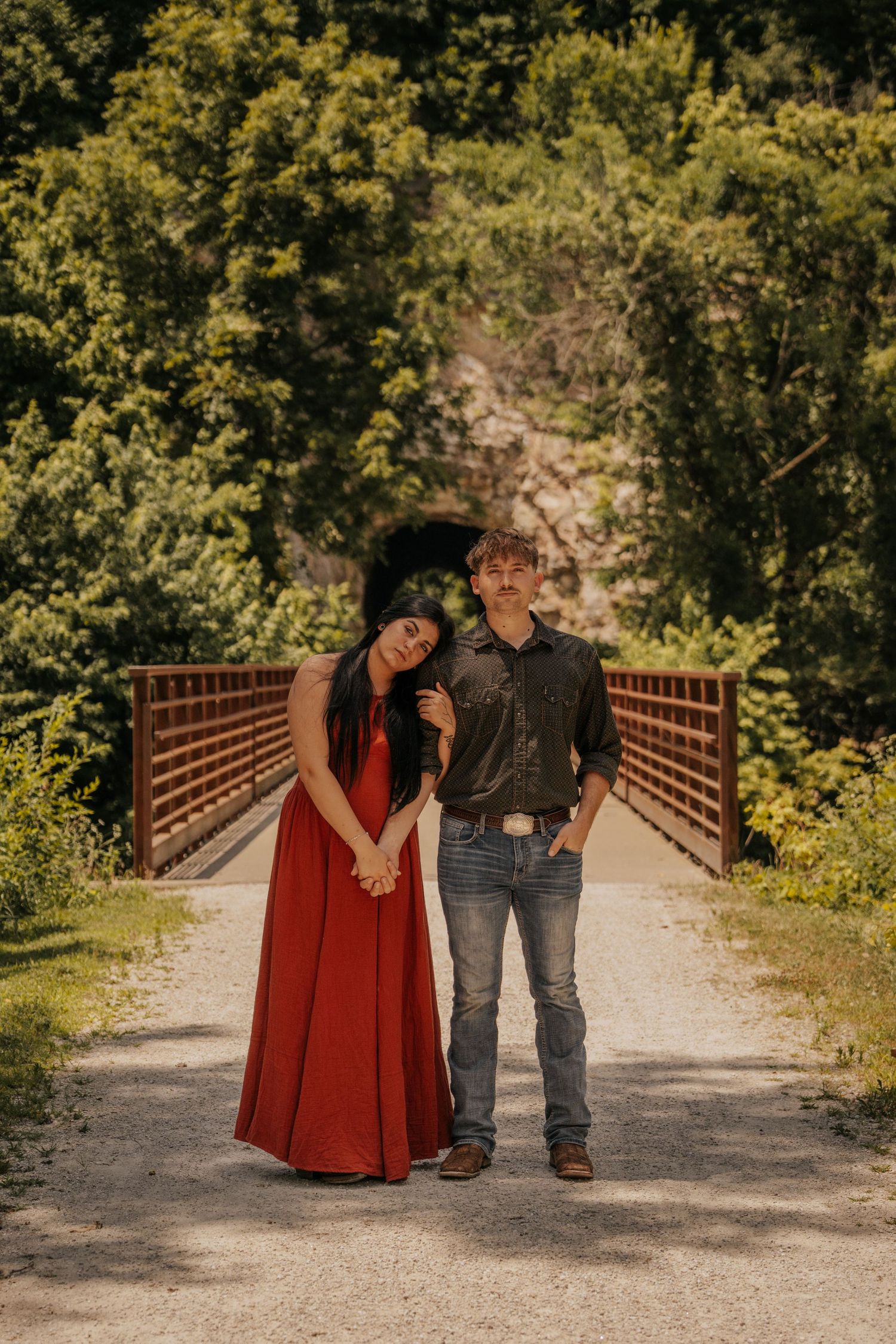 Couple posing on a gravel path by a red bridge, woman in red gown, man in jeans and dark shirt, lush green trees behind.