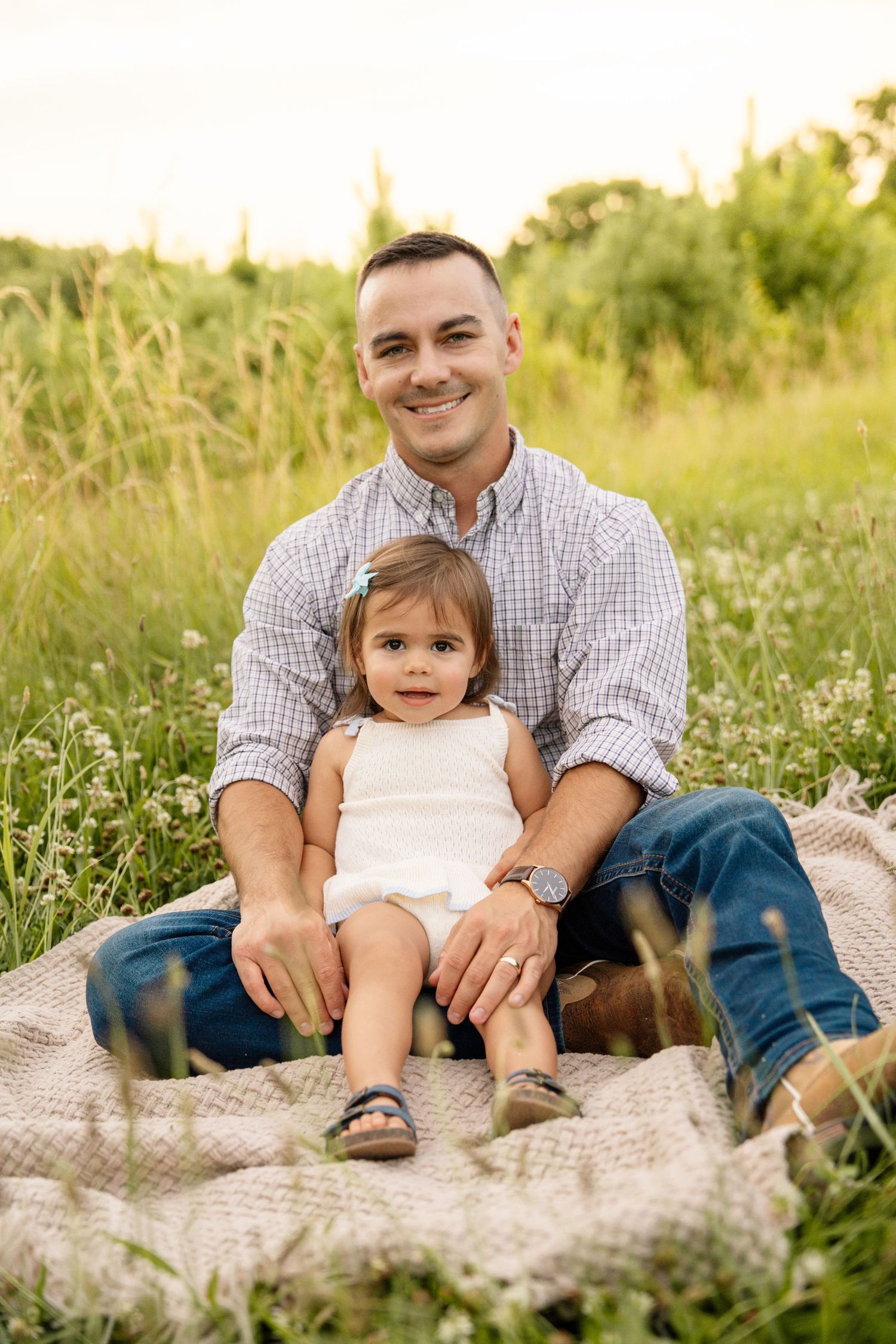 Smiling father sitting on blanket in flower field holding young toddler daughter on his lap during golden hour.