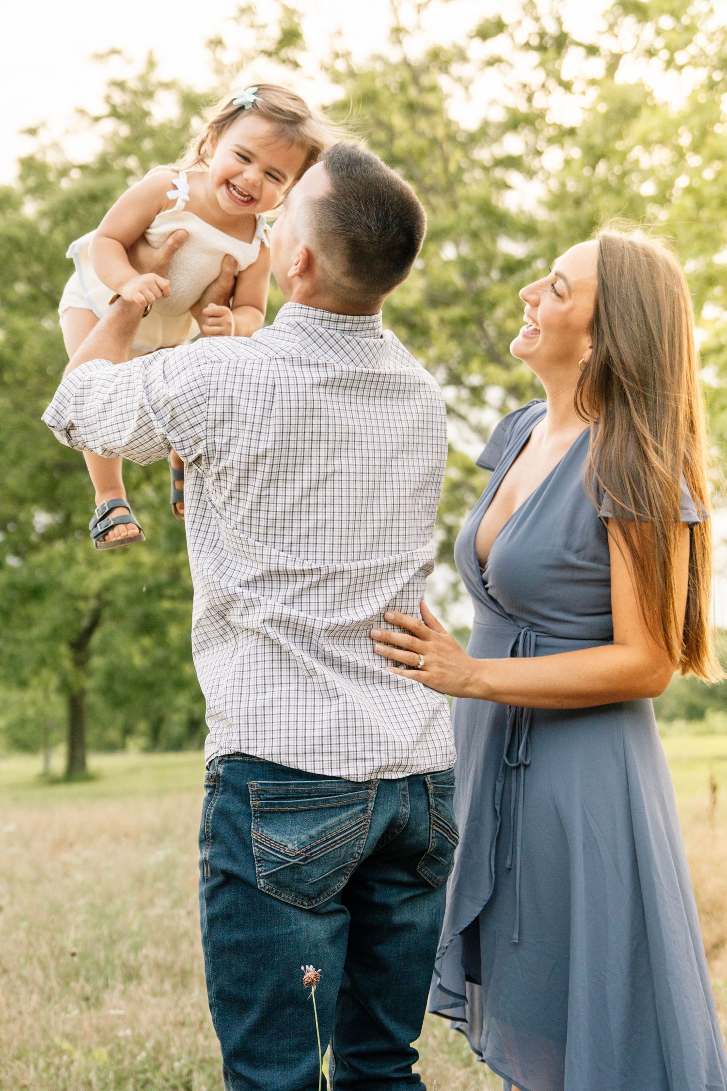 A happy family outdoors, father lifting smiling toddler girl while pregnant mother laughs in a sunny green park.