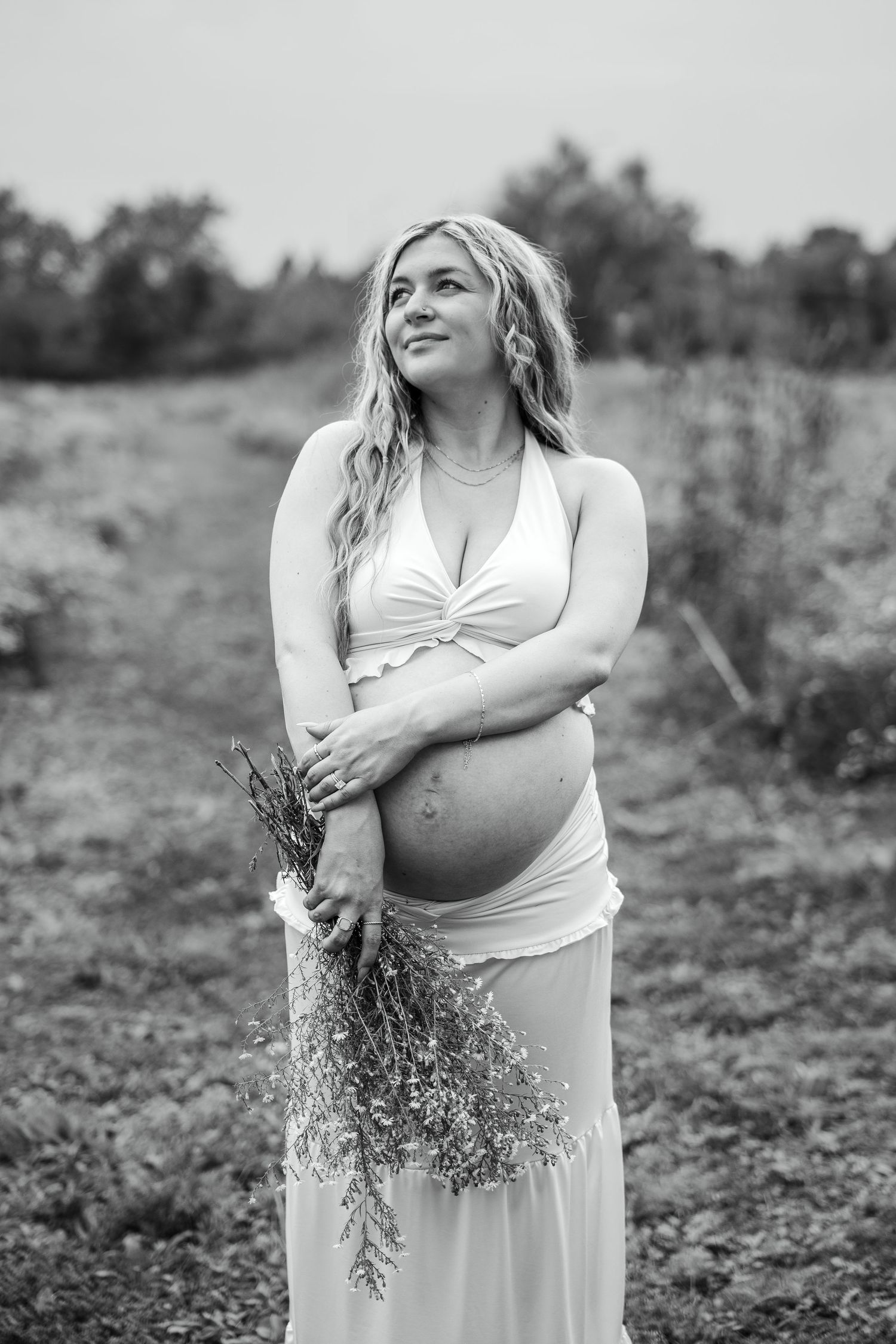 Black and white portrait of smiling pregnant woman holding wildflowers in an open field, wearing white dress.
