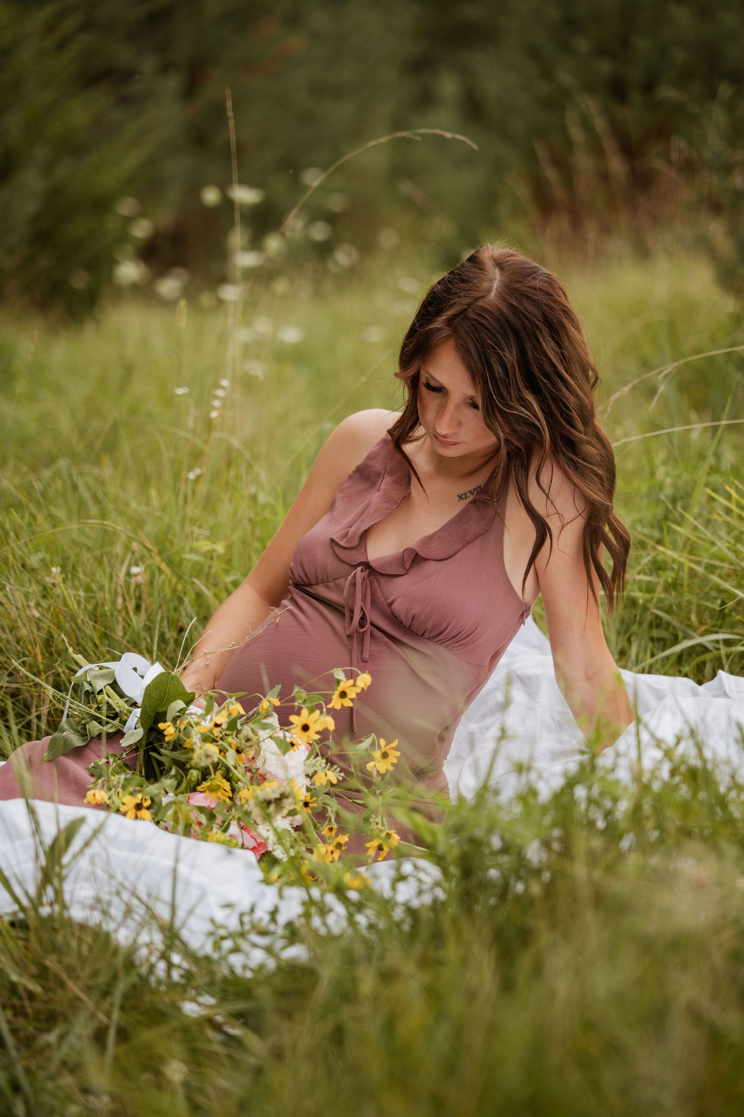 Pregnant woman in mauve dress sits in tall grass with wildflowers bouquet during outdoor maternity photo session.