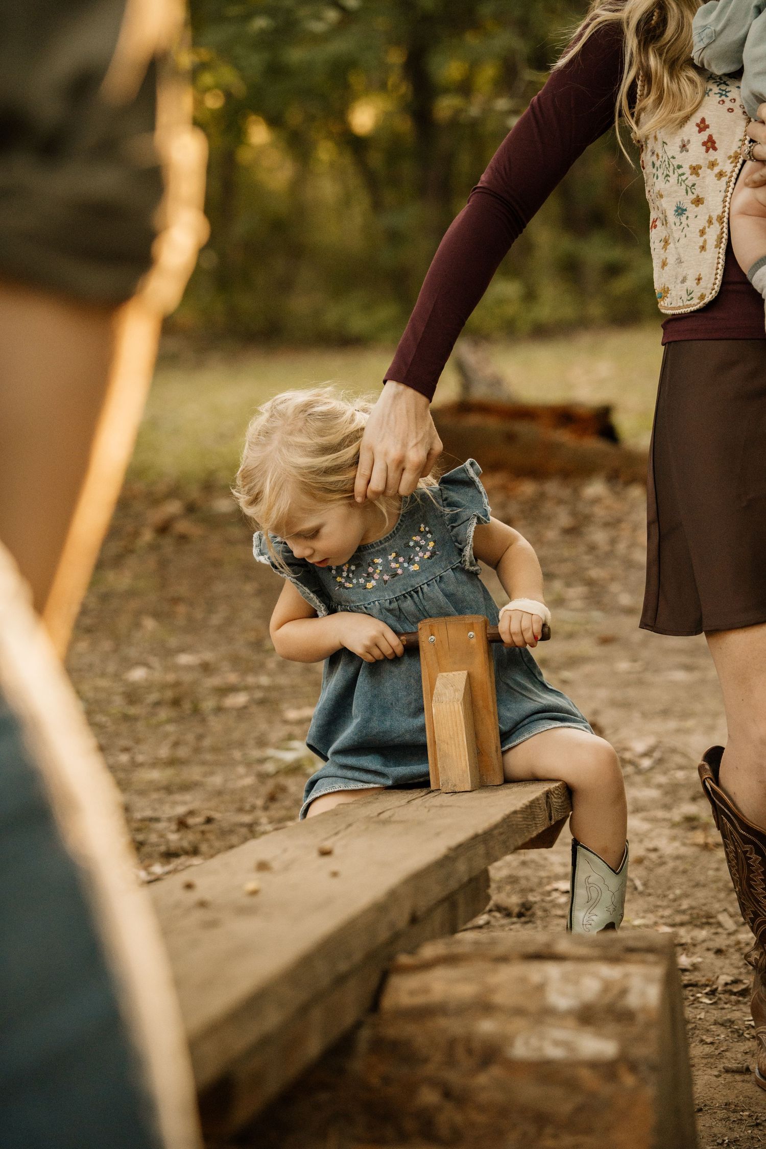 A toddler girl in a blue dress plays on a log bench in a woodland setting.