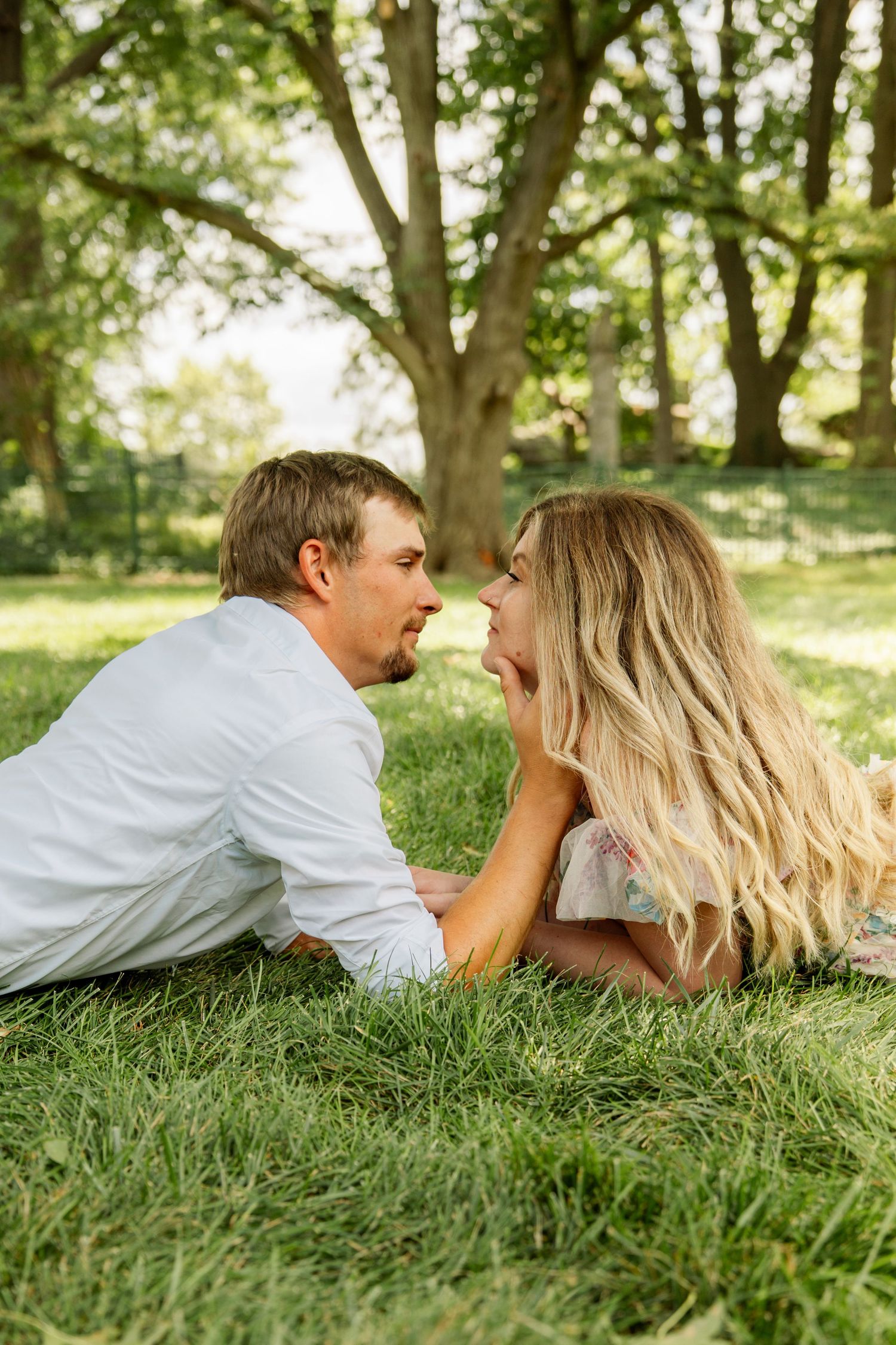 Romantic couple lying on green grass face to face beneath large trees in a sunny park during an outdoor photo session.