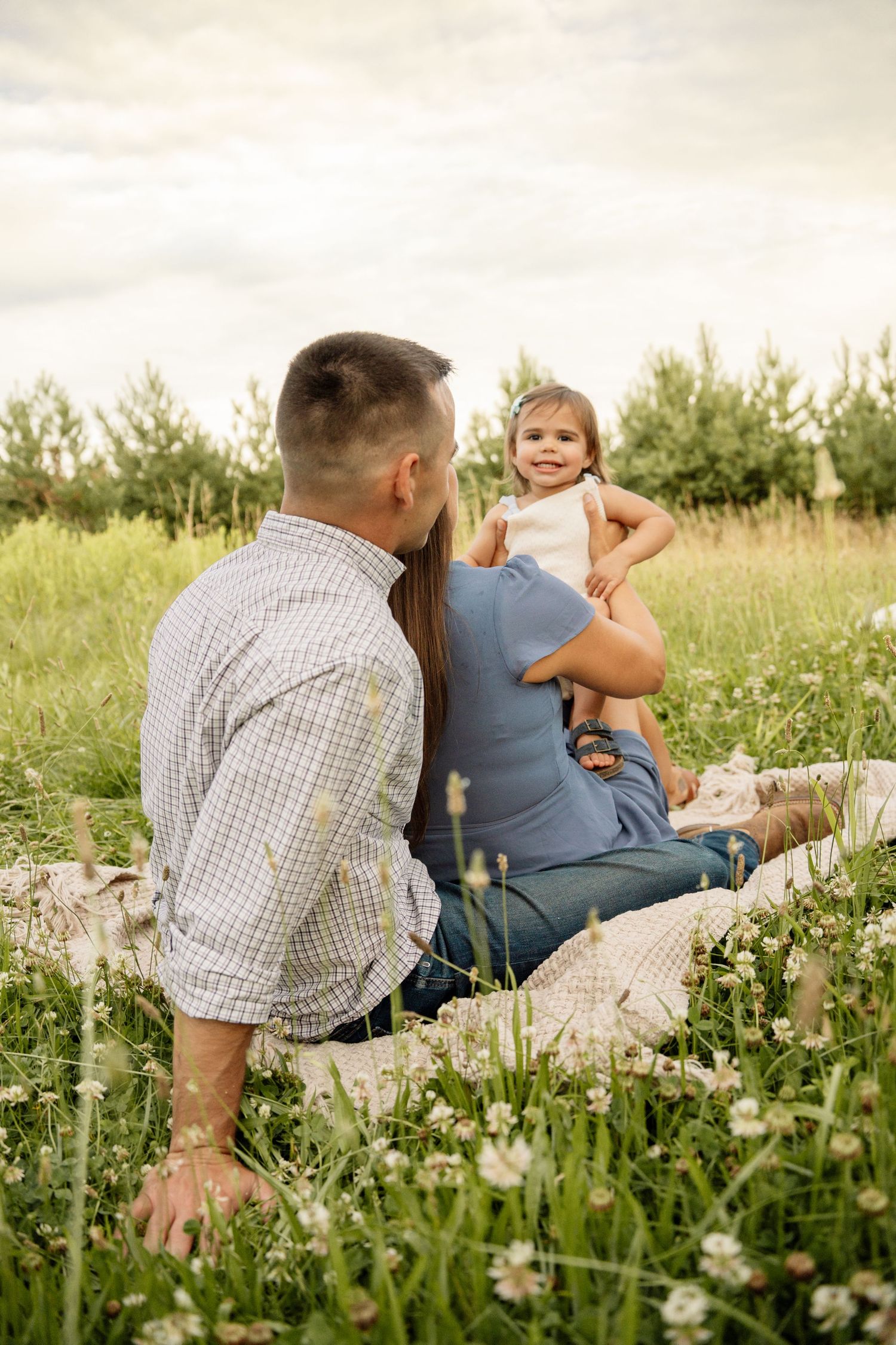 Family sitting in a wildflower field, parents facing toddler girl who smiles joyfully on a sunny day.
