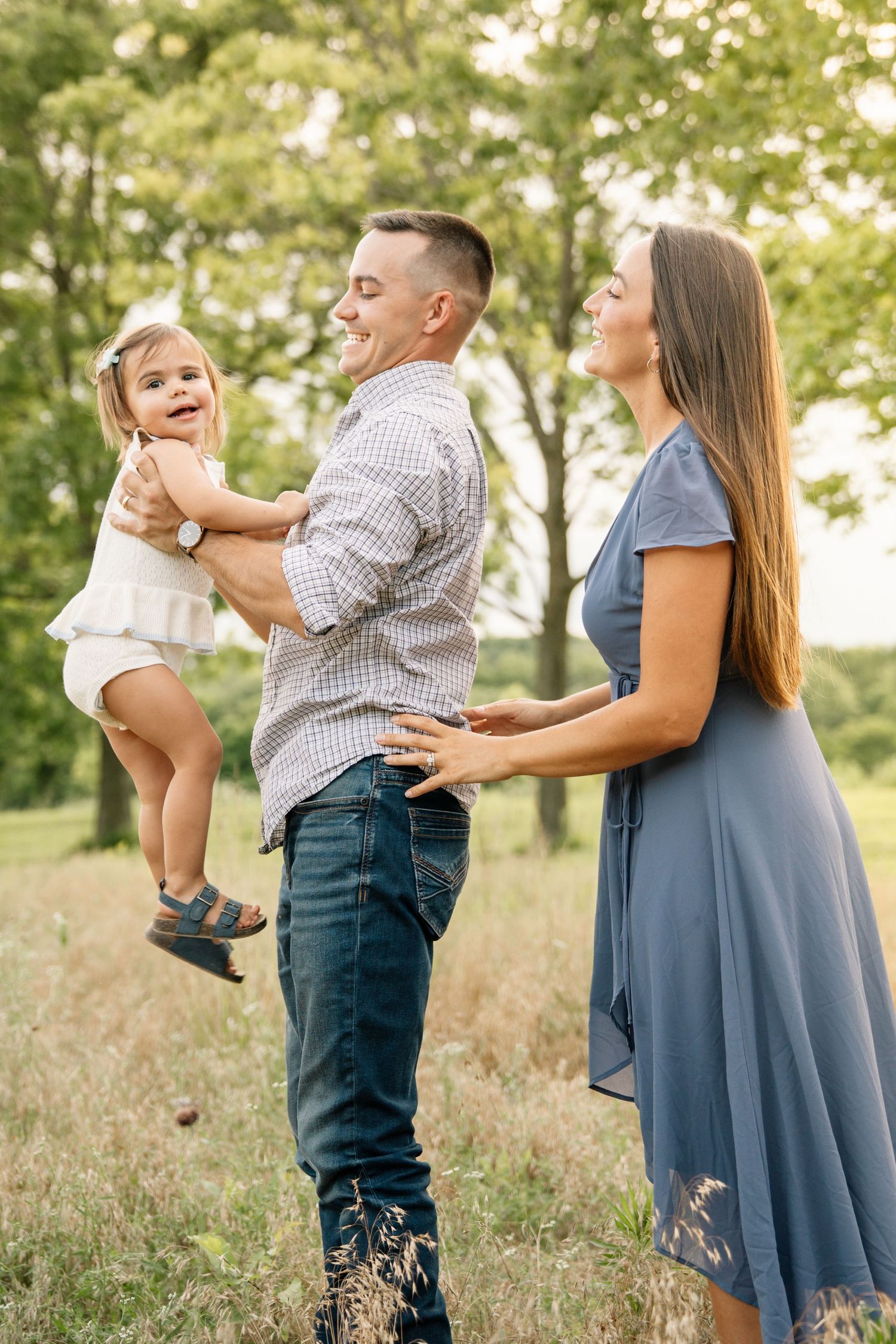 Happy family with dad holding toddler girl while mom smiles in sunny green park.