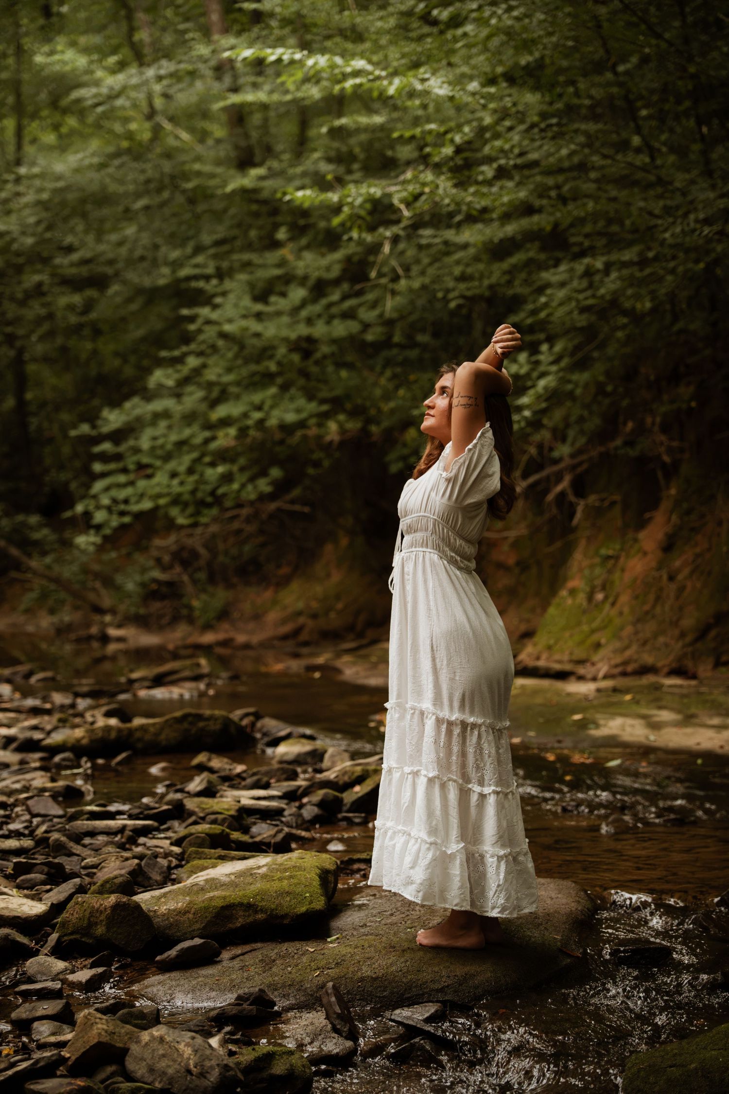 Young woman in flowing white dress stands barefoot on creek stones in lush green forest.