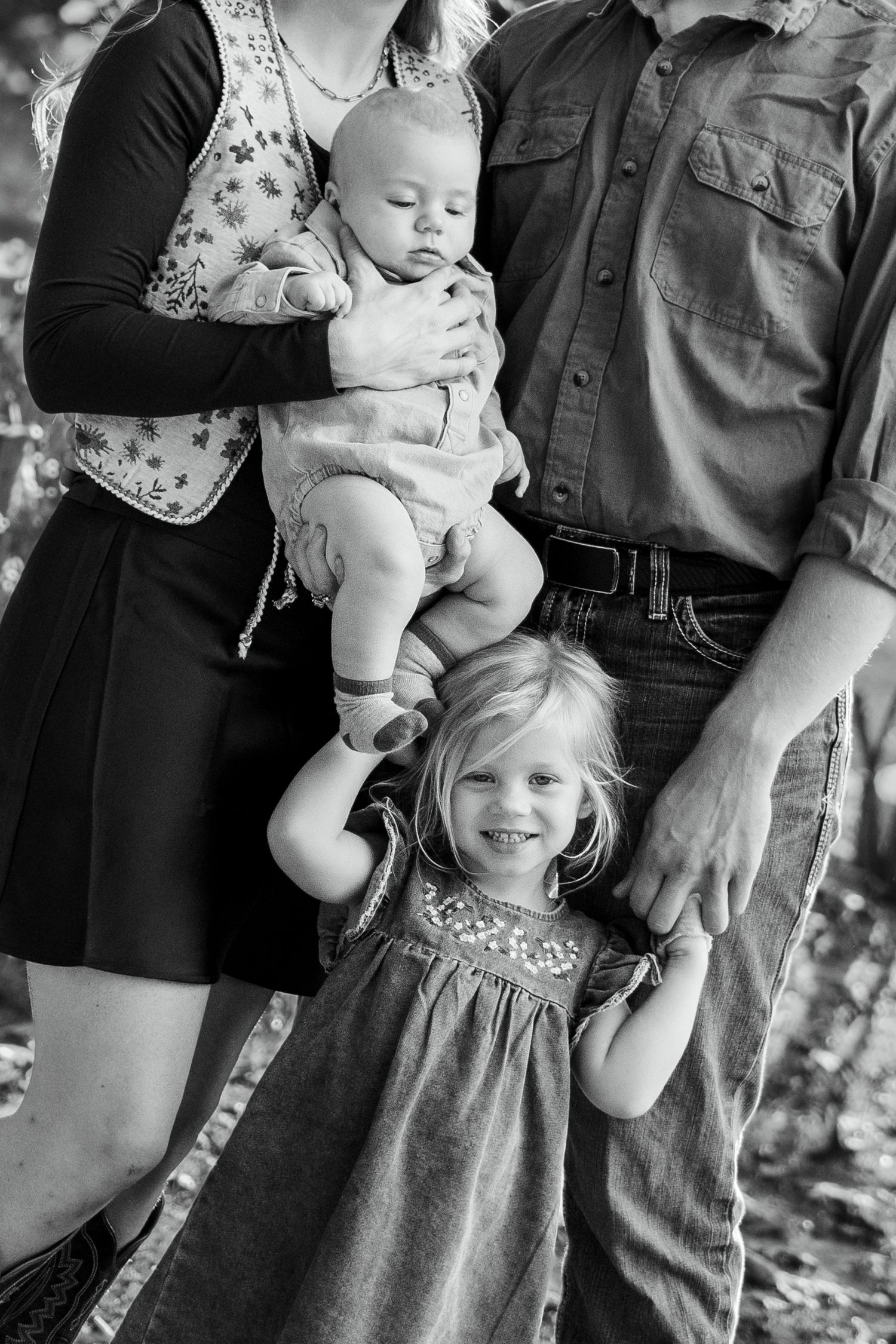 Black and white family photo with parents holding a baby and a smiling toddler girl standing between them outdoors.