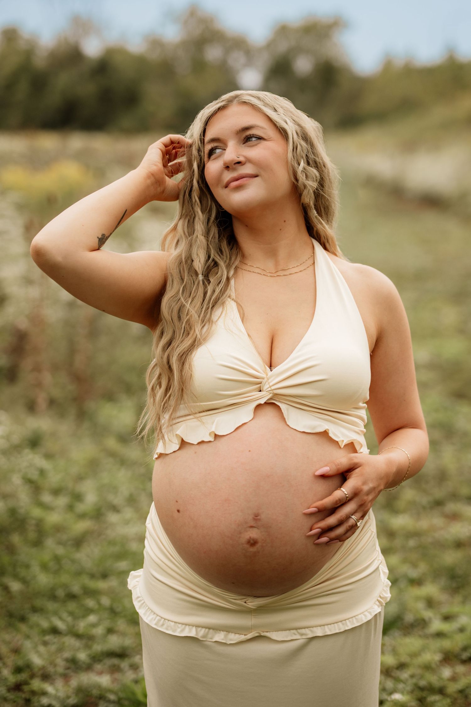 Smiling pregnant woman in cream halter top cradling baby bump outdoors in natural setting.