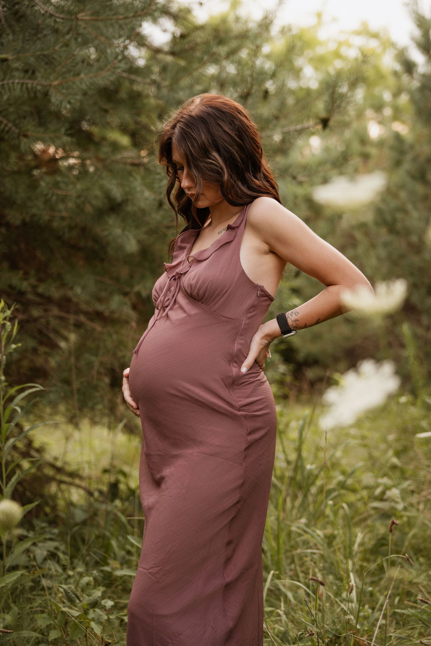 Pregnant woman in mauve sleeveless dress stands among lush greenery with hand on hip outdoors.