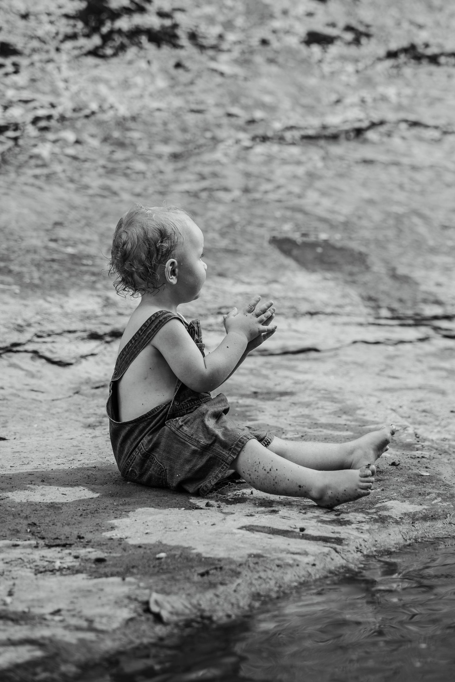 Black and white photo of a baby in overalls sitting by a rocky riverbank.
