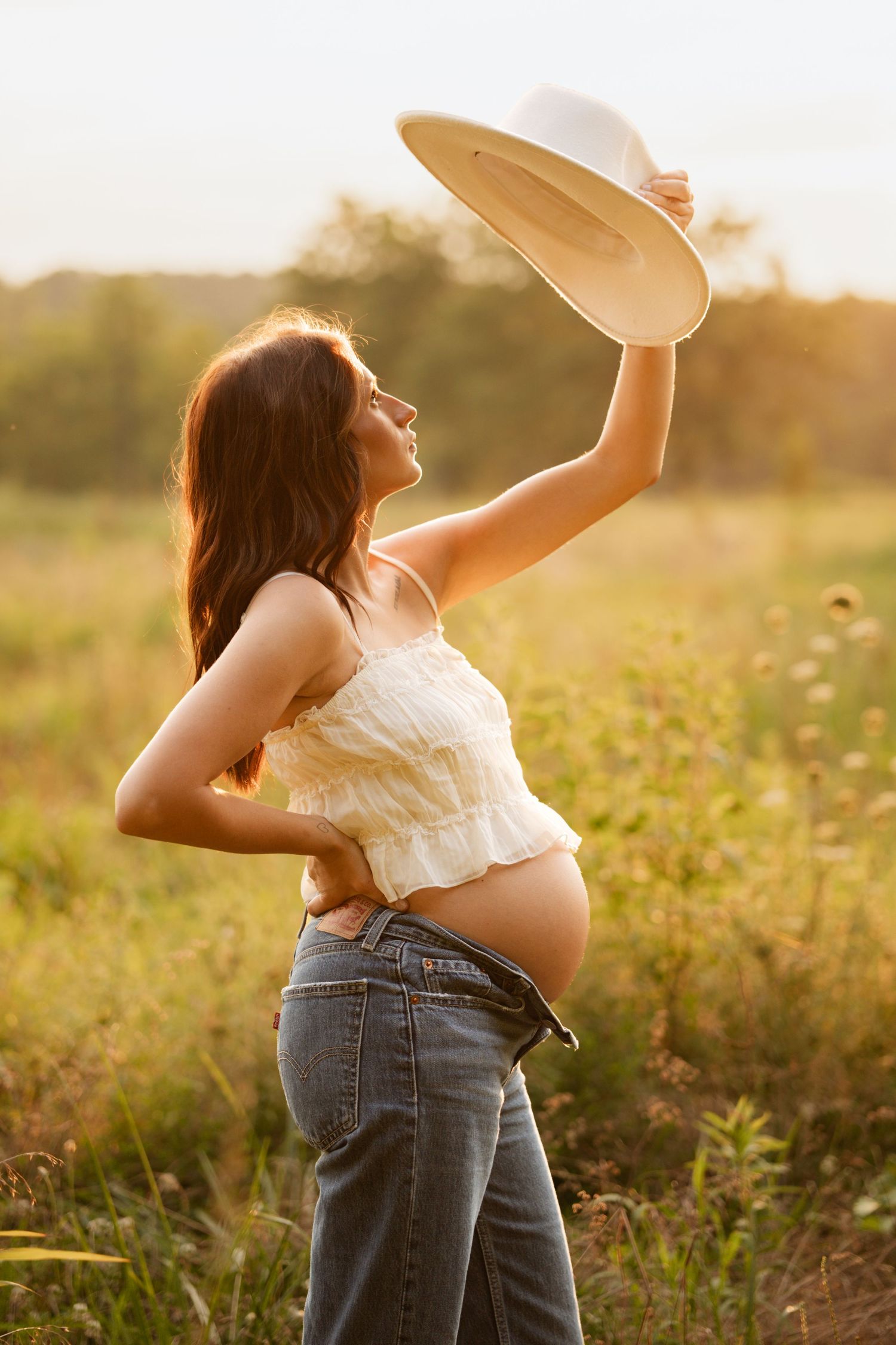 Pregnant woman in white crop top raising a hat in a golden sunset meadow.