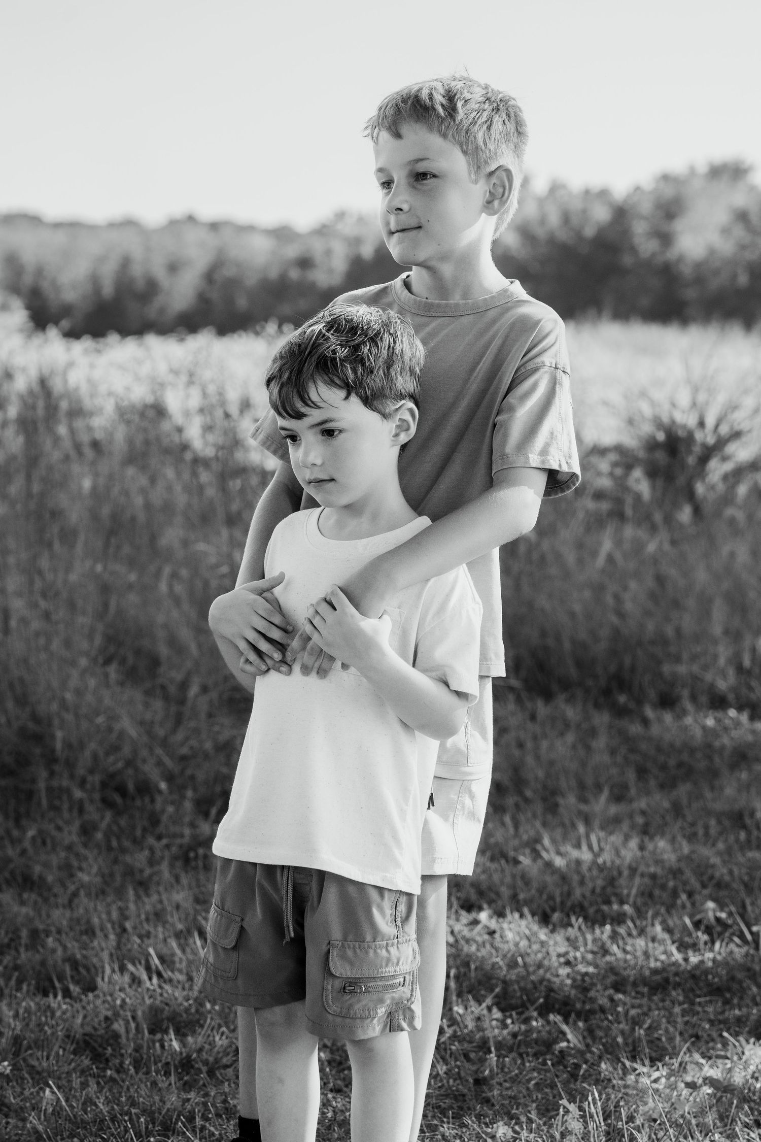 Two boys embracing outdoors in a black and white photo, older brother hugging younger sibling in a grassy field.