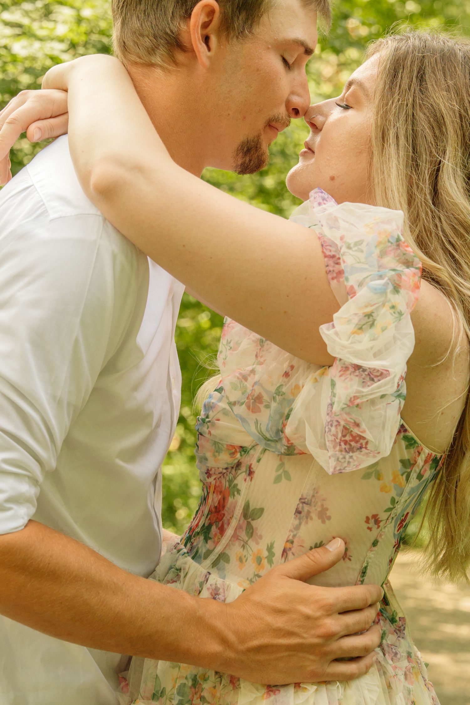 Couple leaning in for a kiss outdoors, woman in floral off-shoulder dress embracing man in white shirt.