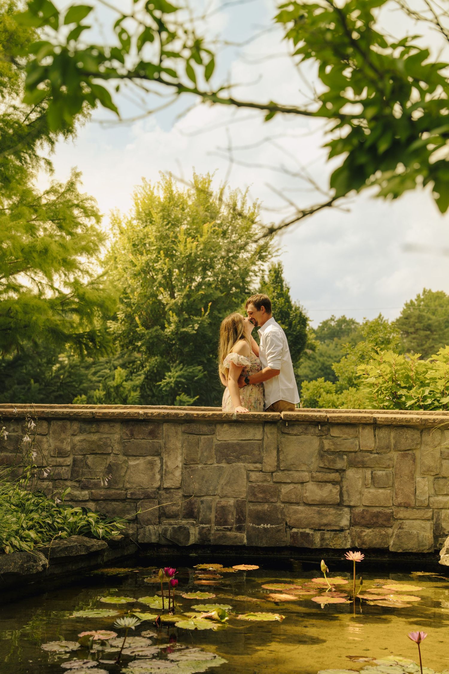 Couple sharing a romantic kiss on a stone bridge over a lily pond, surrounded by lush green gardens in golden sunlight.
