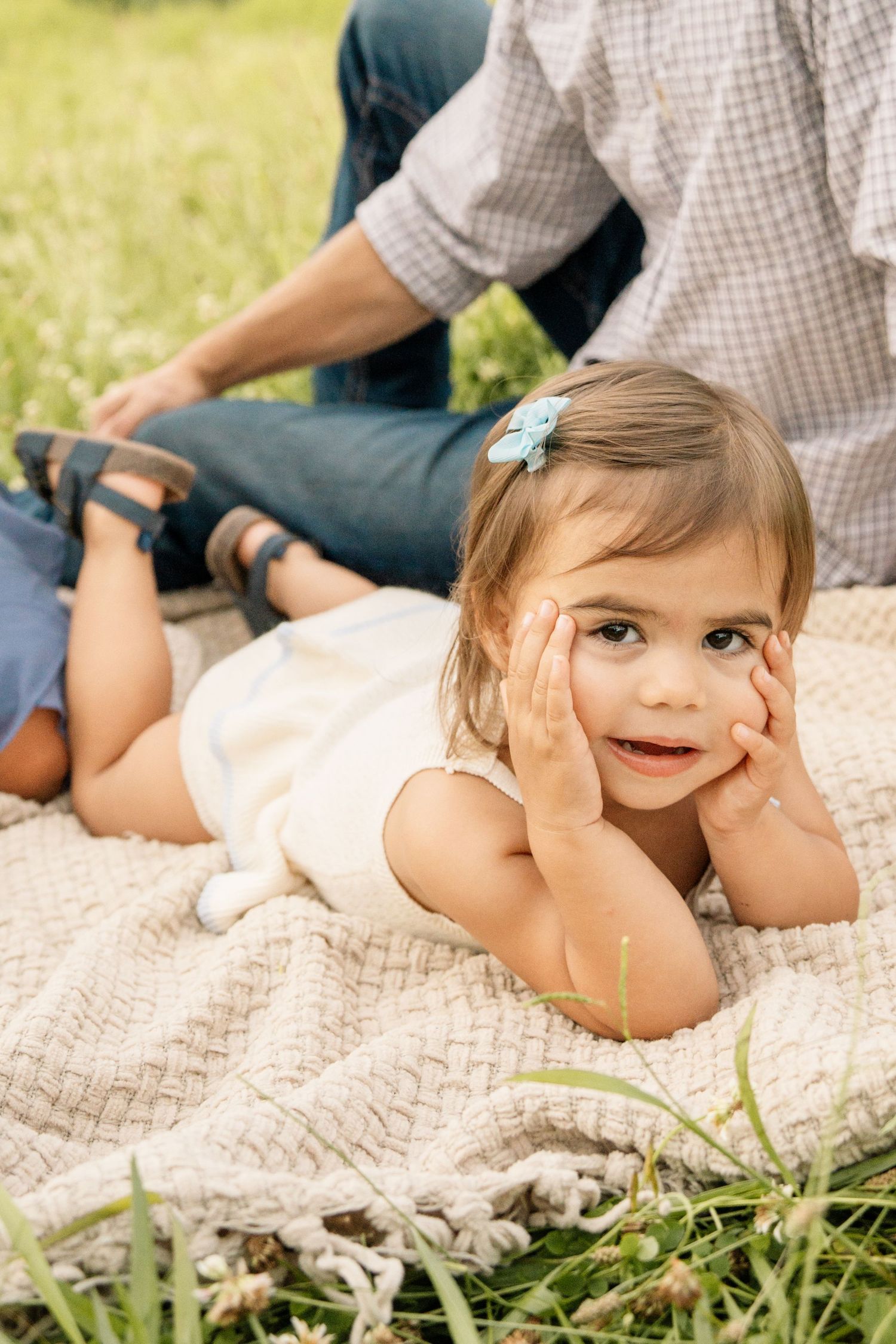 Cute toddler girl with blue bow lying on blanket in meadow resting chin on hands.