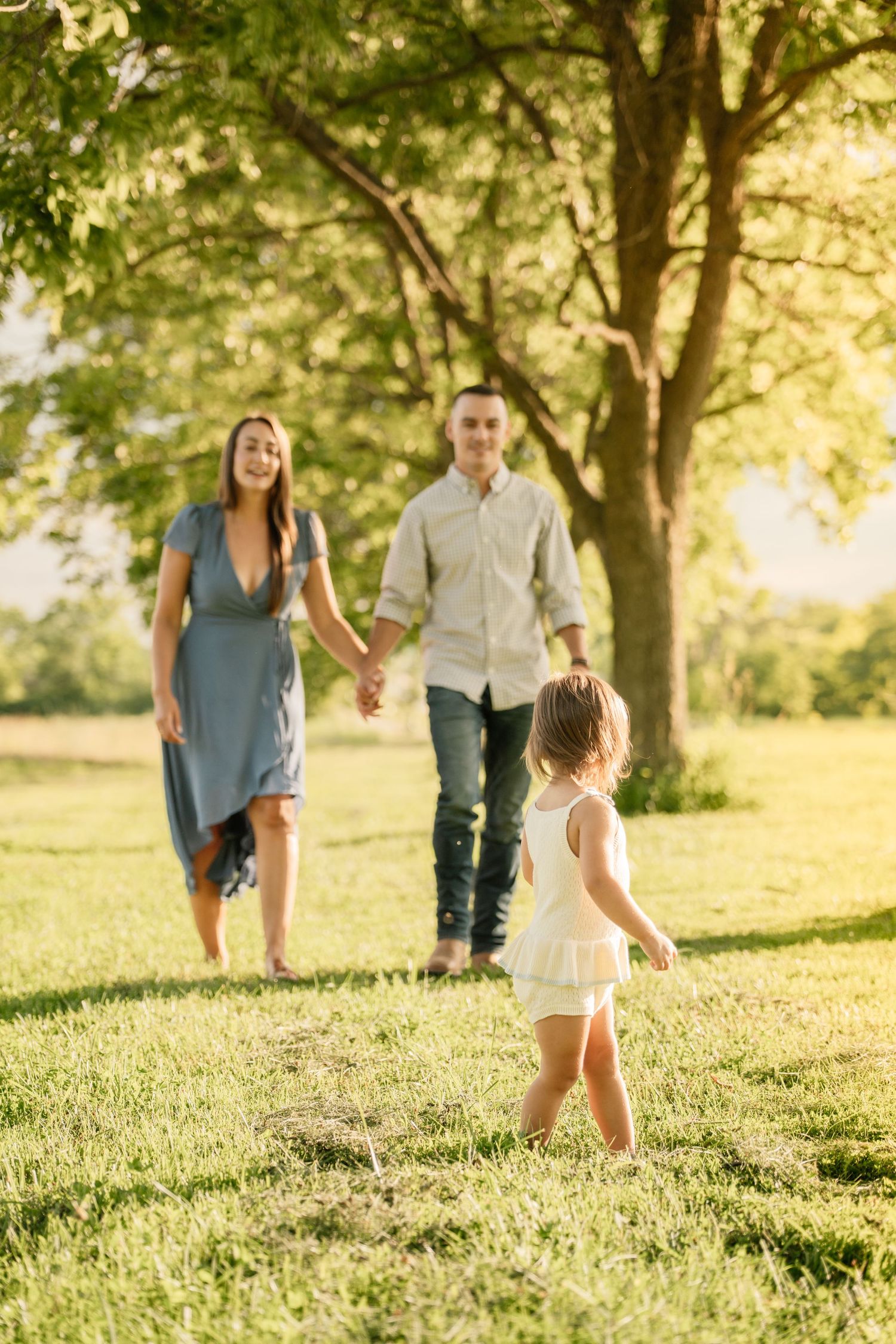 Family holding hands in sunny park, young girl in white dress walks ahead of smiling parents outdoors.