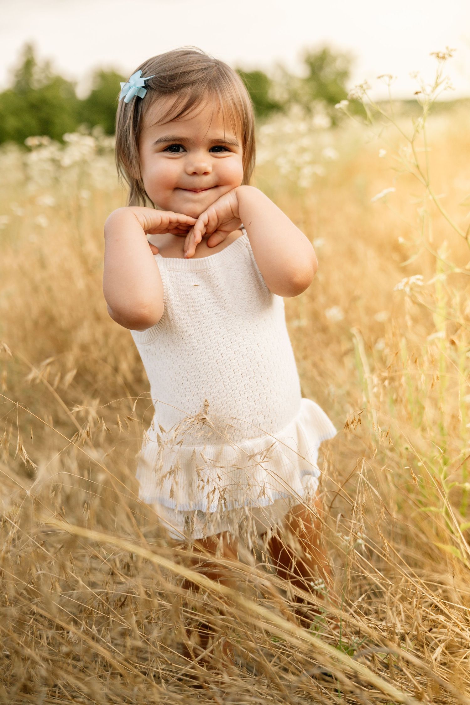 Smiling toddler girl in white dress with blue hair bow standing in golden field.