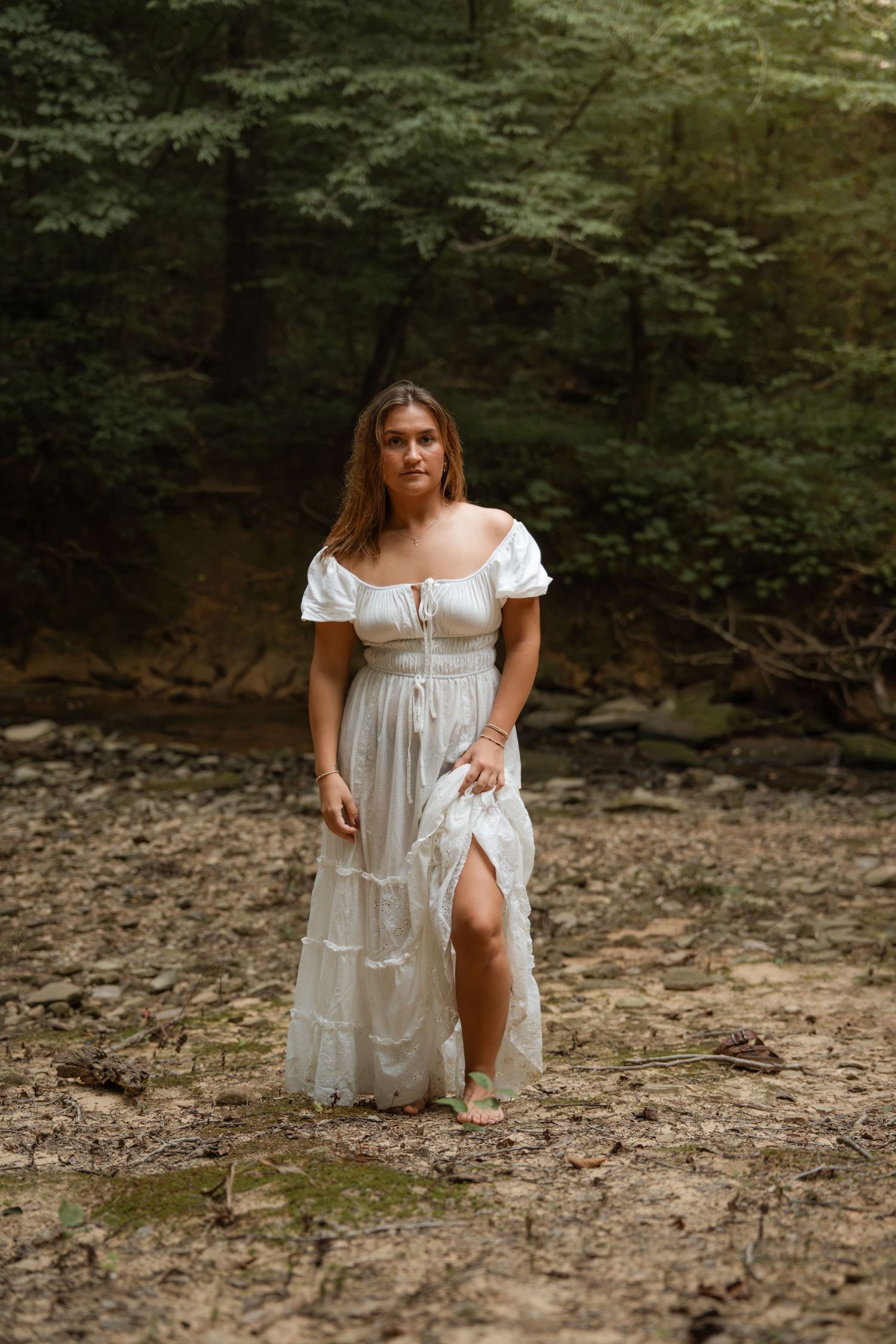 Young woman in white off-shoulder maxi dress posing in a lush forest setting.