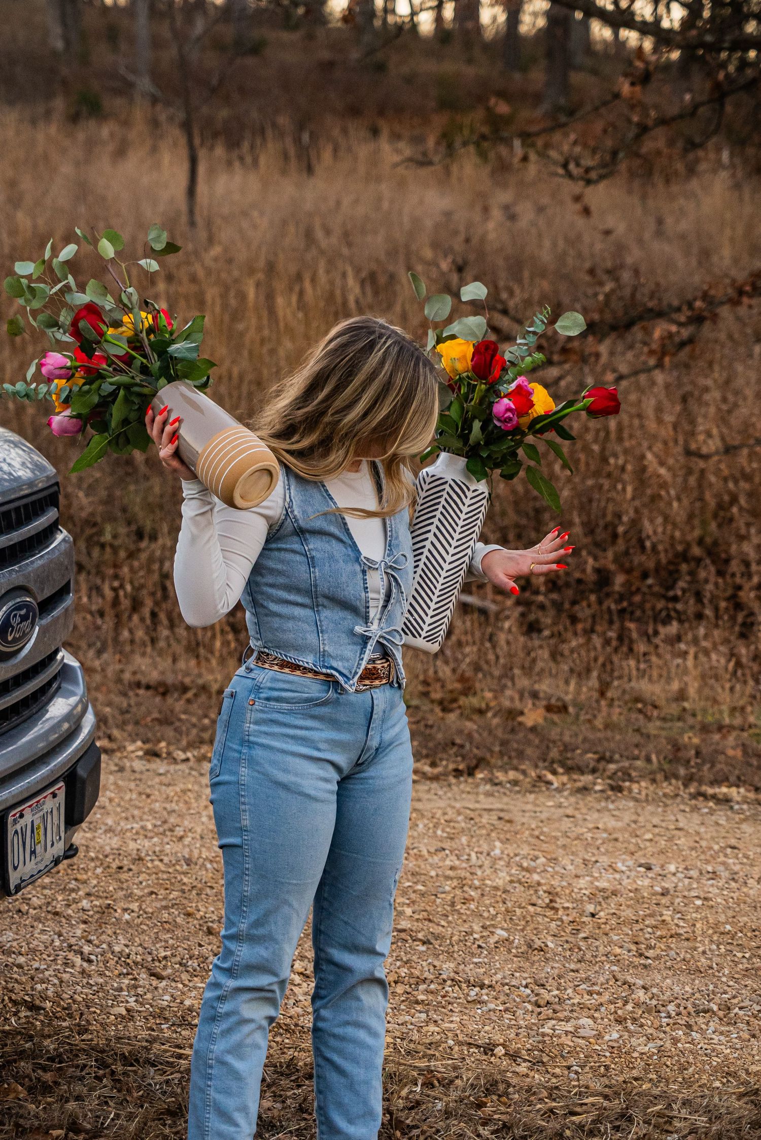Woman in denim overalls carrying two colorful floral arrangements near a truck in a rural setting.