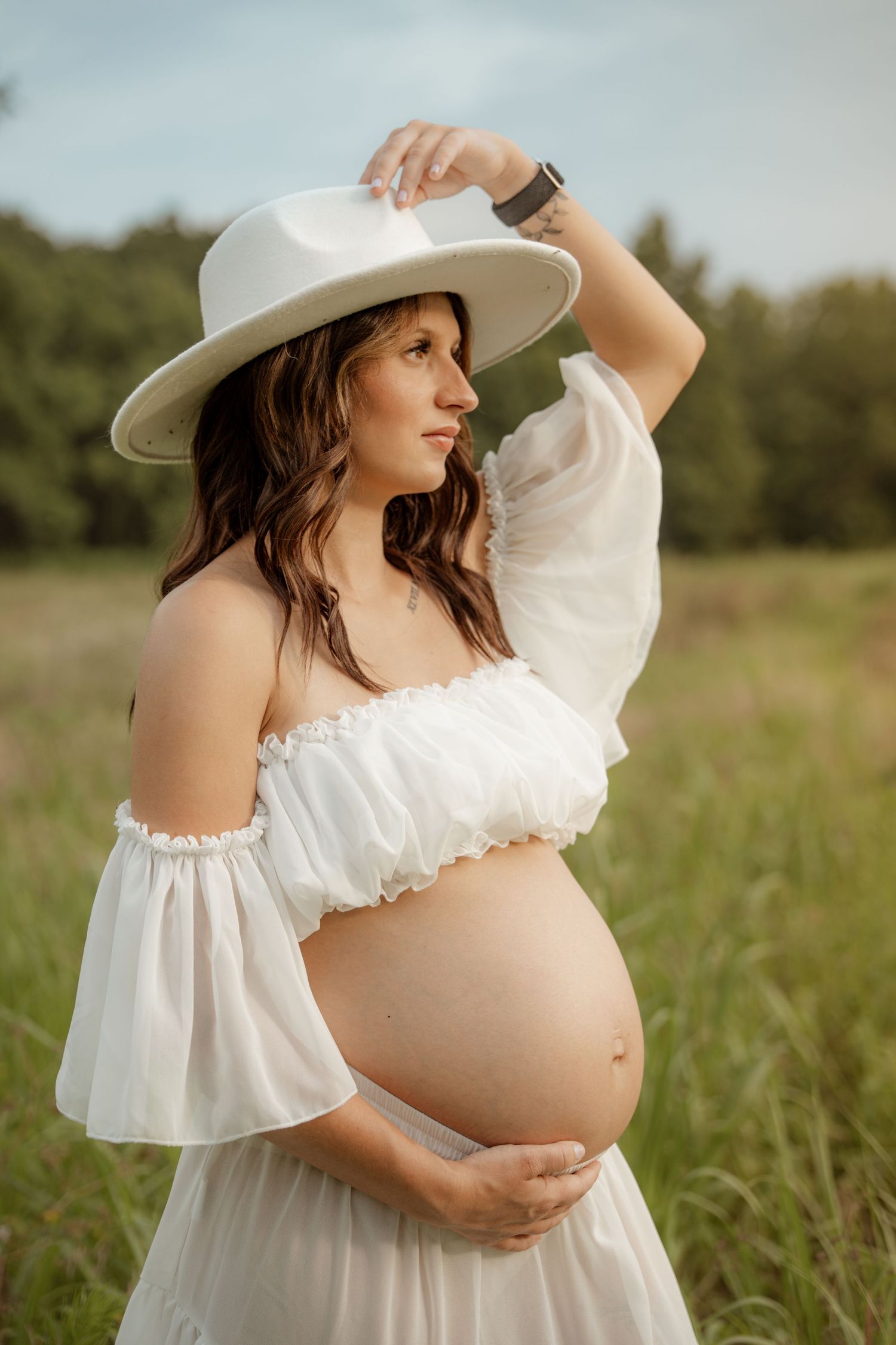 Side profile of pregnant woman in white ruffled crop top and hat cradling baby bump in lush outdoor setting.