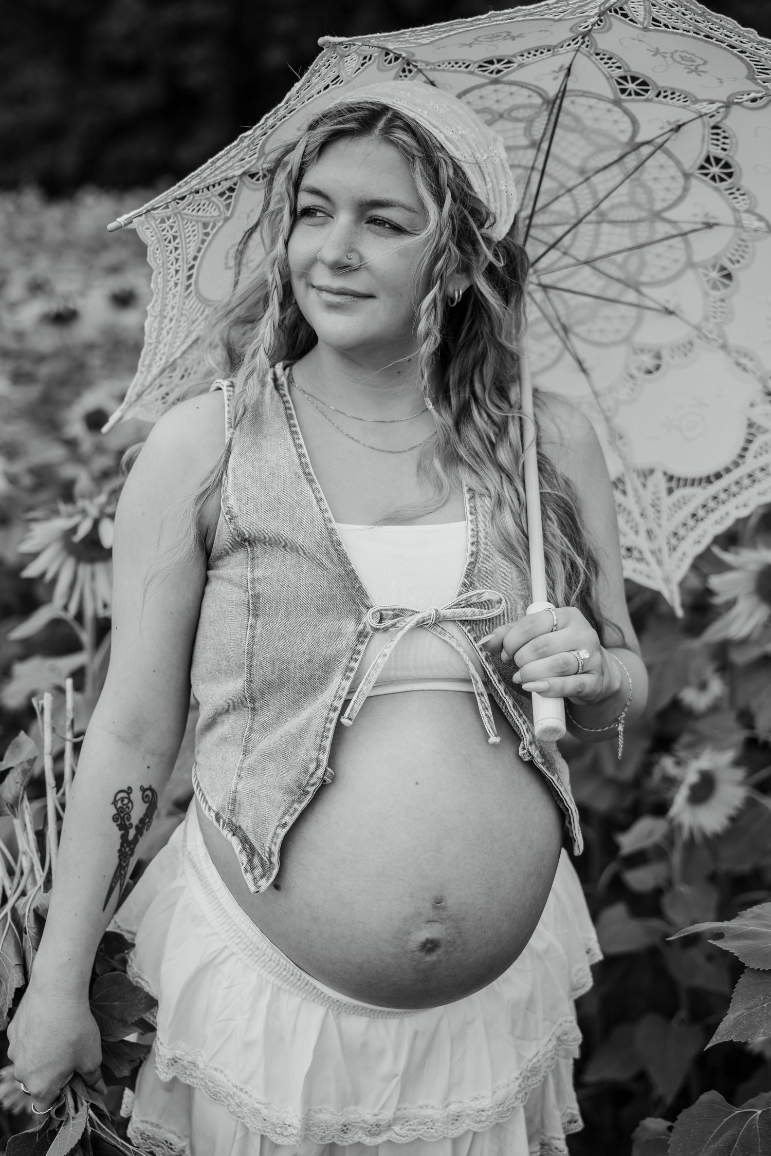 Pregnant woman holding lace parasol in sunflower field, wearing denim vest and white skirt, black and white photo.