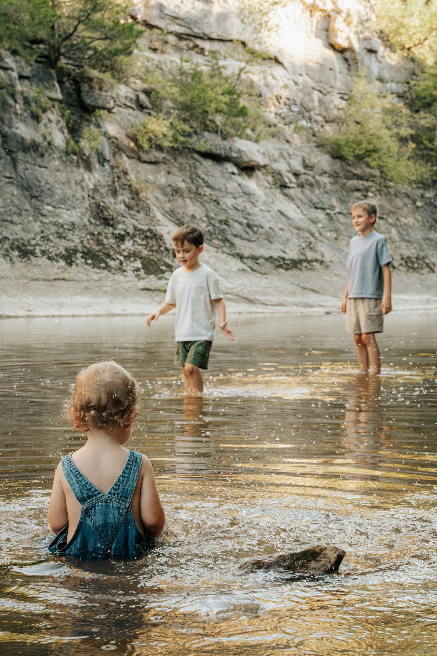 Three children playing in a shallow river near rocky cliffs, with a toddler girl sitting in the water.