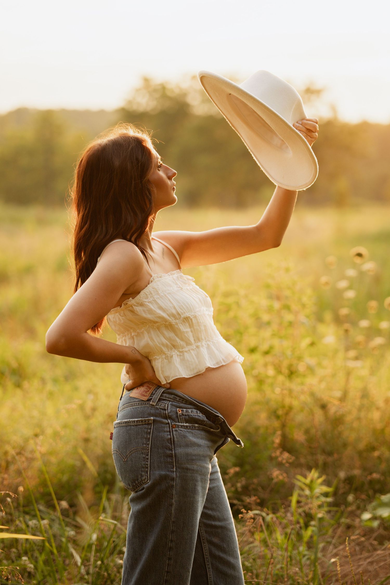 Pregnant woman in white crop top and jeans holds hat up in sunny field at golden hour.