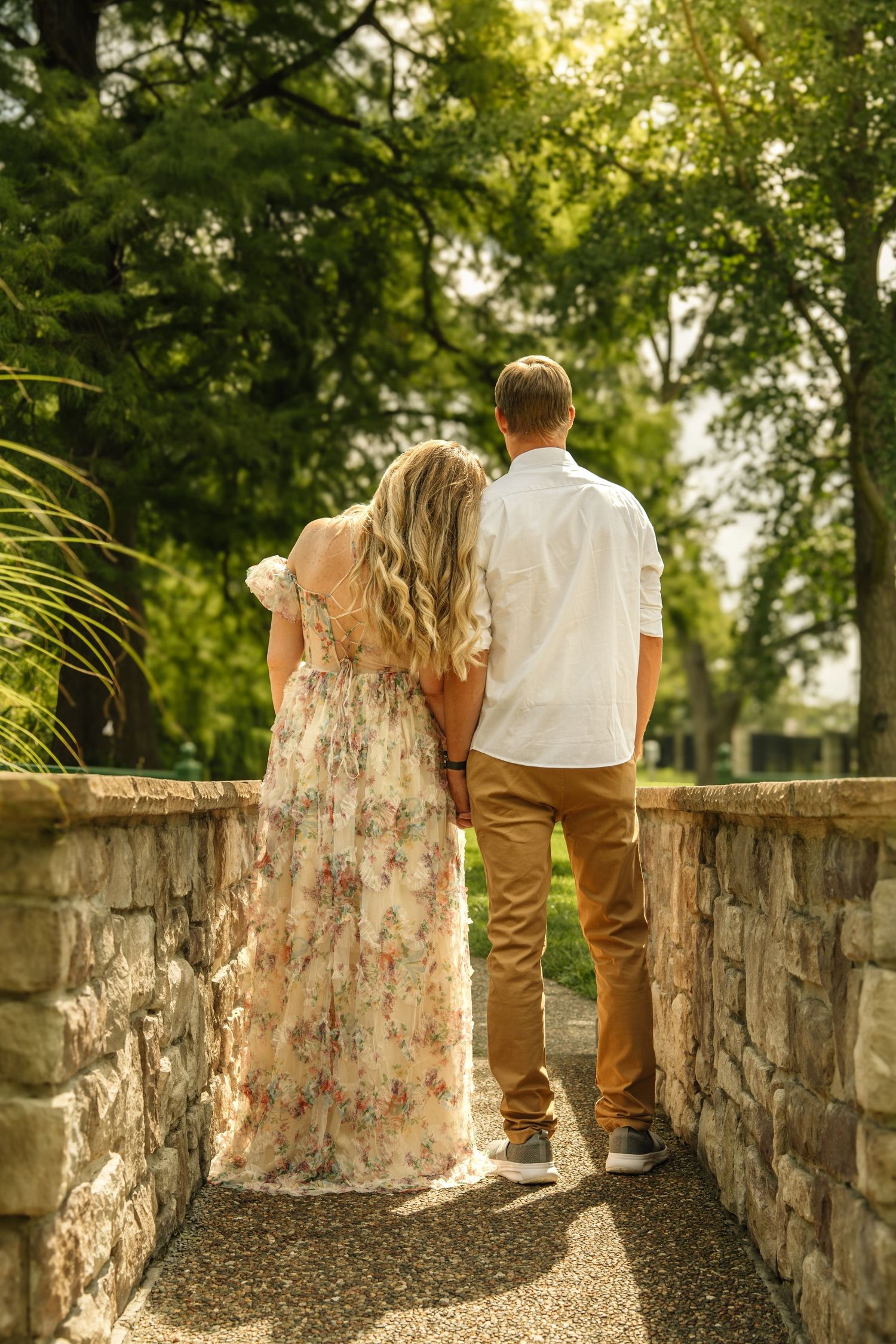 Couple holding hands from behind on a stone bridge pathway surrounded by sunlit green trees.