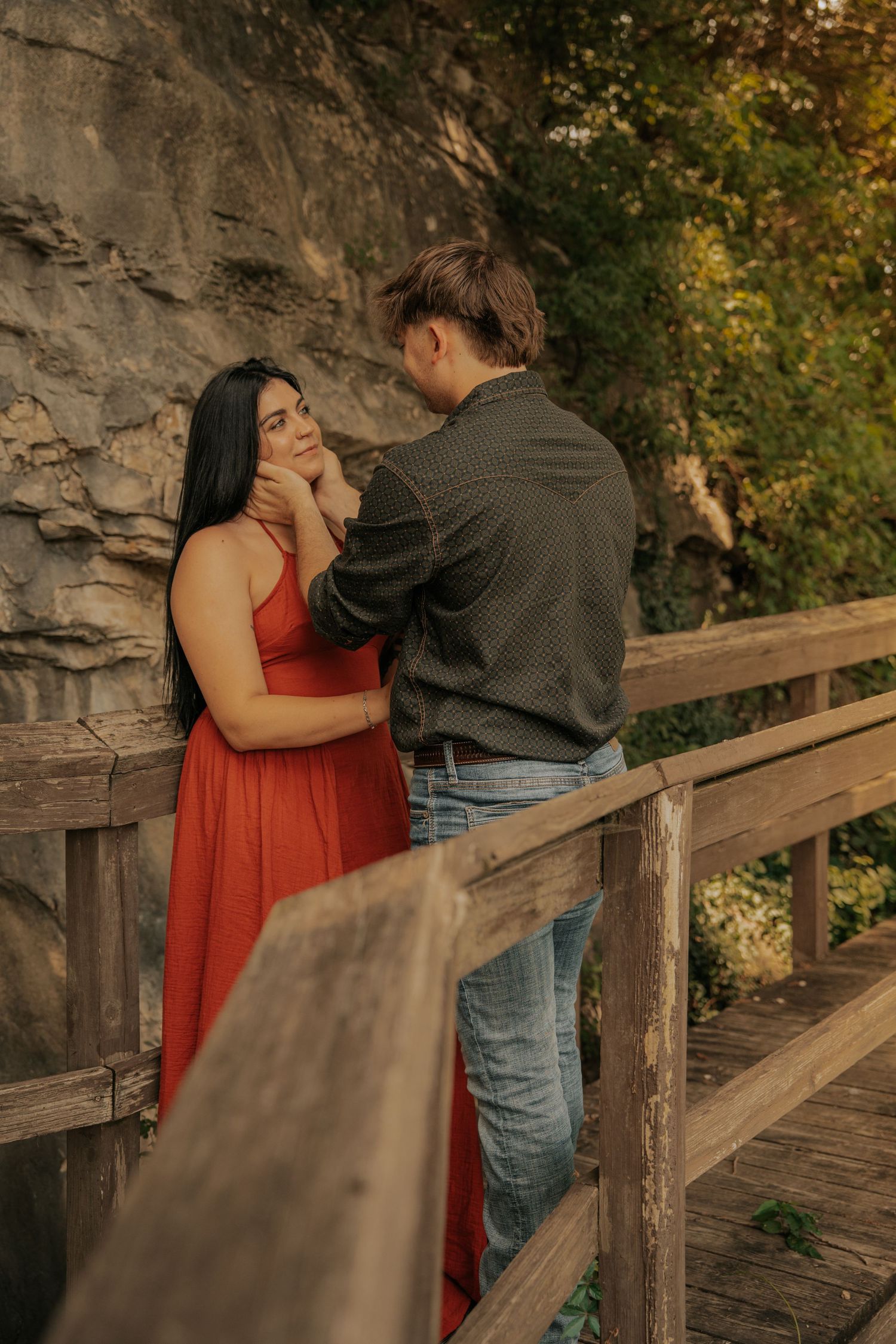 Couple sharing a romantic moment on a wooden bridge, woman in red dress, man in gray shirt, with rocky cliff backdrop.