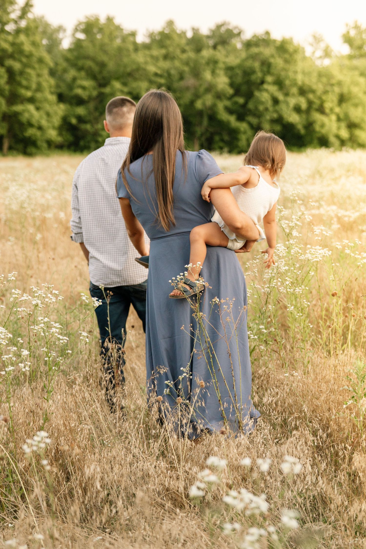 Family of three walking through a sunny wildflower meadow, mother in blue dress carrying child.