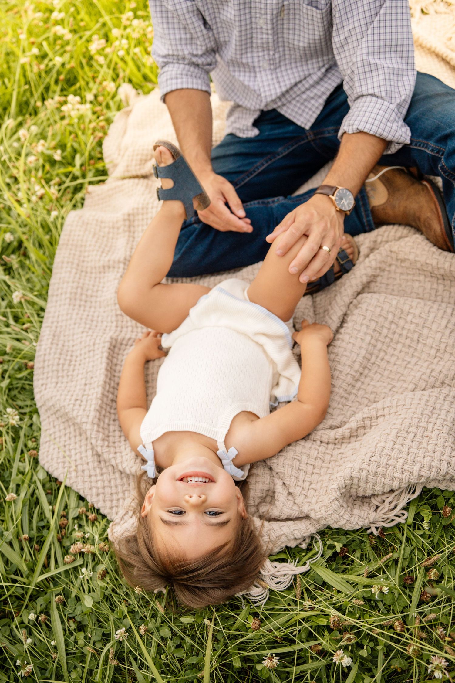 Overhead view of a toddler girl lying on a blanket in grass, looking up while her father plays with her.