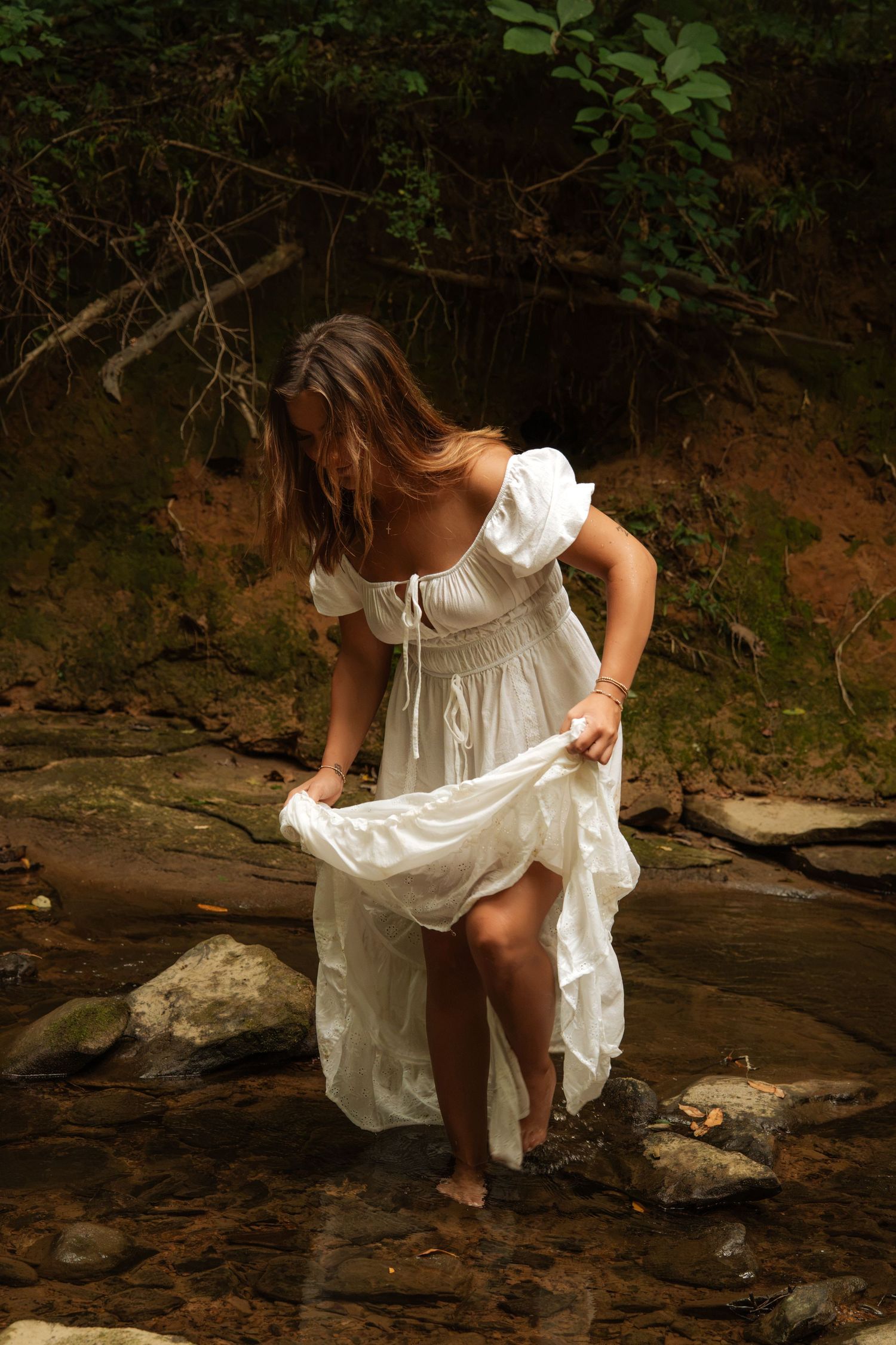 Woman in white off-shoulder dress lifting skirt while wading barefoot on rocky creek in lush forest.