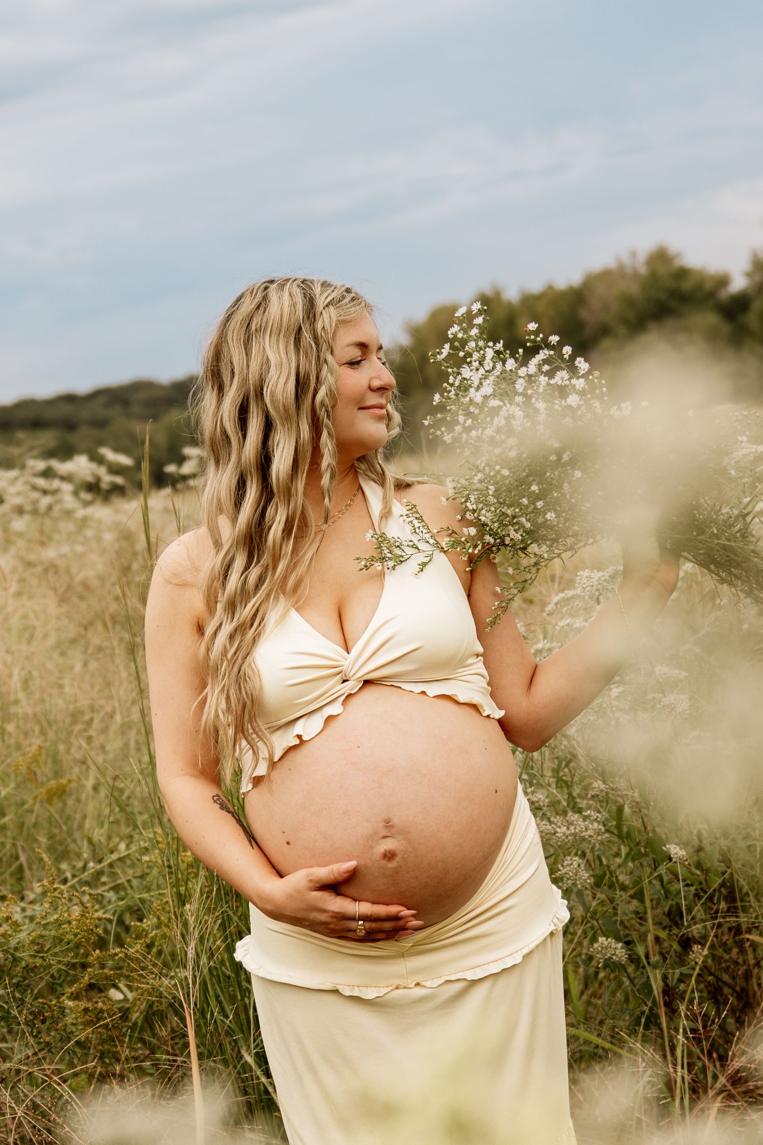 Pregnant woman in cream halter dress holding wildflowers in a meadow, smiling serenely outdoors.
