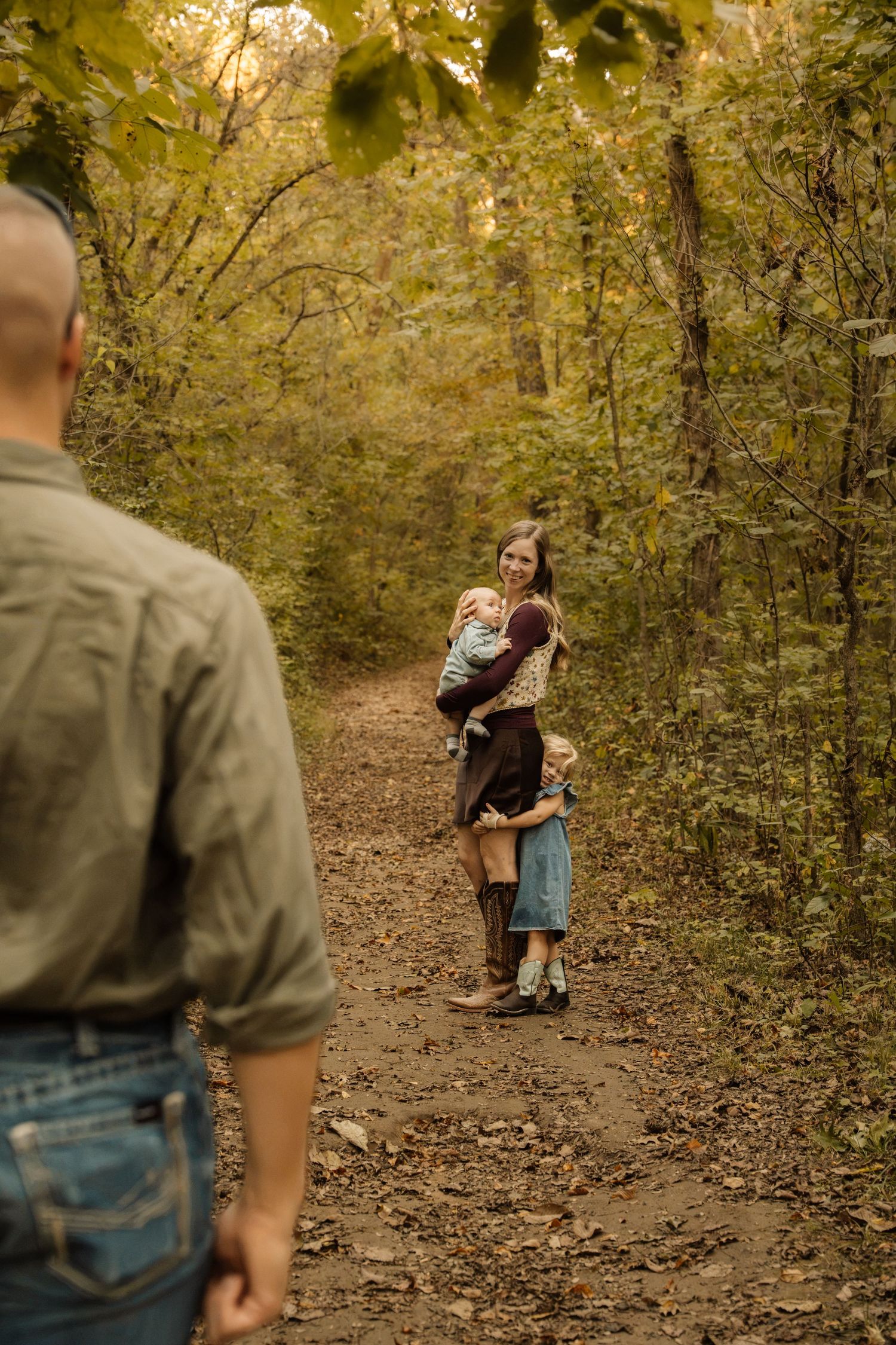 Family walking on a wooded autumn trail, mother holding toddler with young child beside her as father approaches.