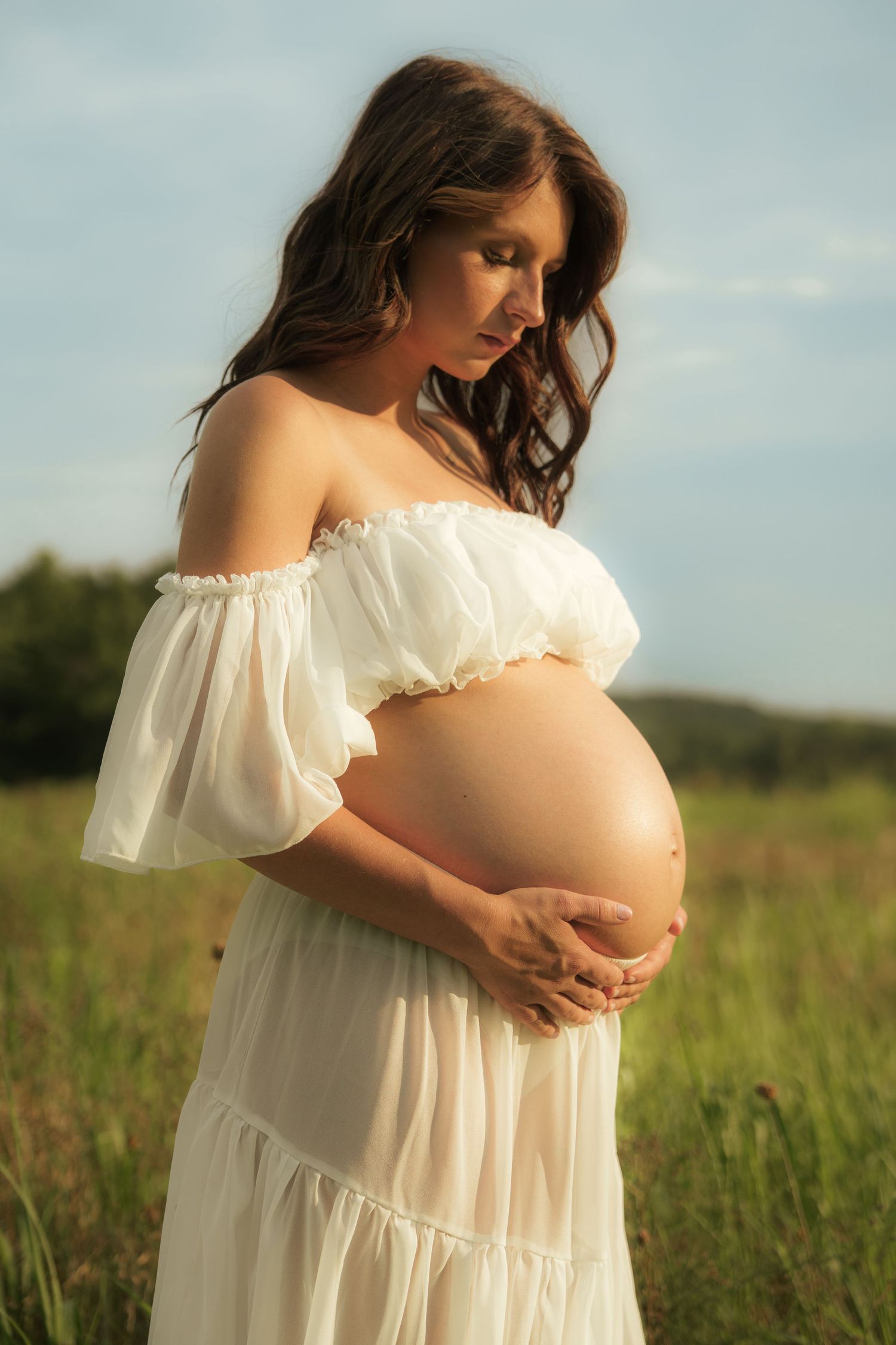 Pregnant woman in white off-shoulder dress cradling her baby bump in a sunny green field.