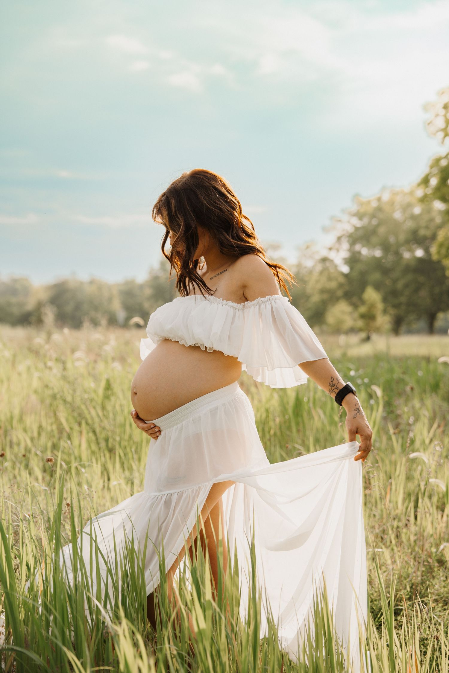 Pregnant woman in white off-shoulder dress turns away showing baby bump while holding flowing skirt in field.