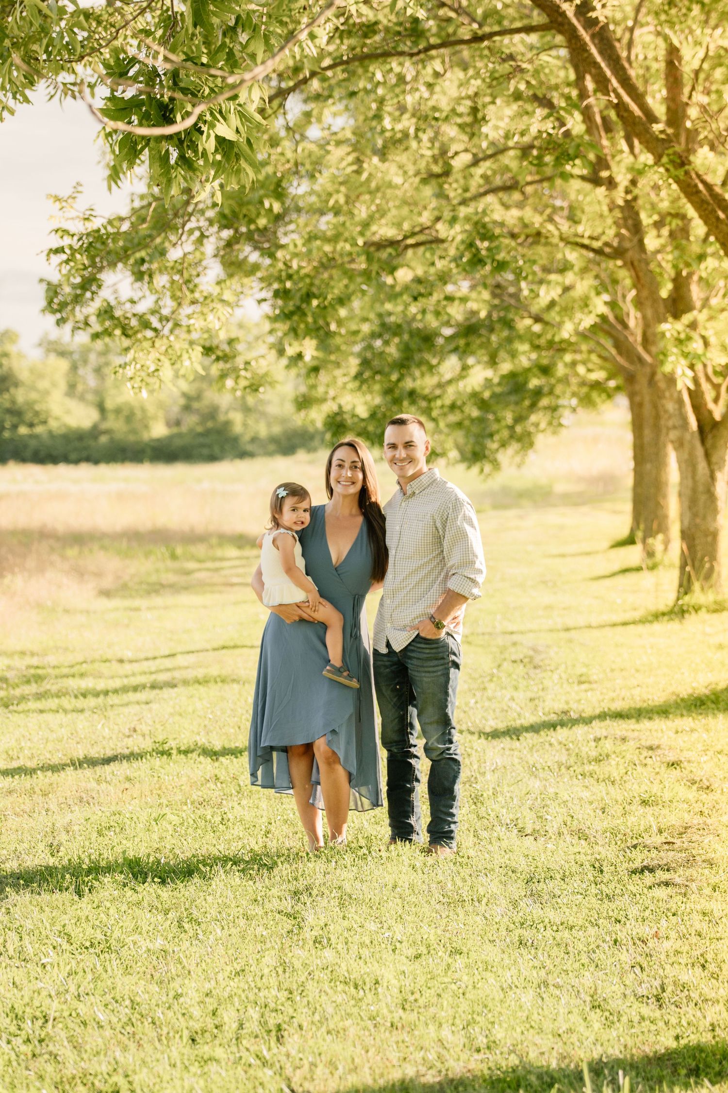 A happy young family with a toddler poses under a tree in a sunny green field, mother in blue dress.