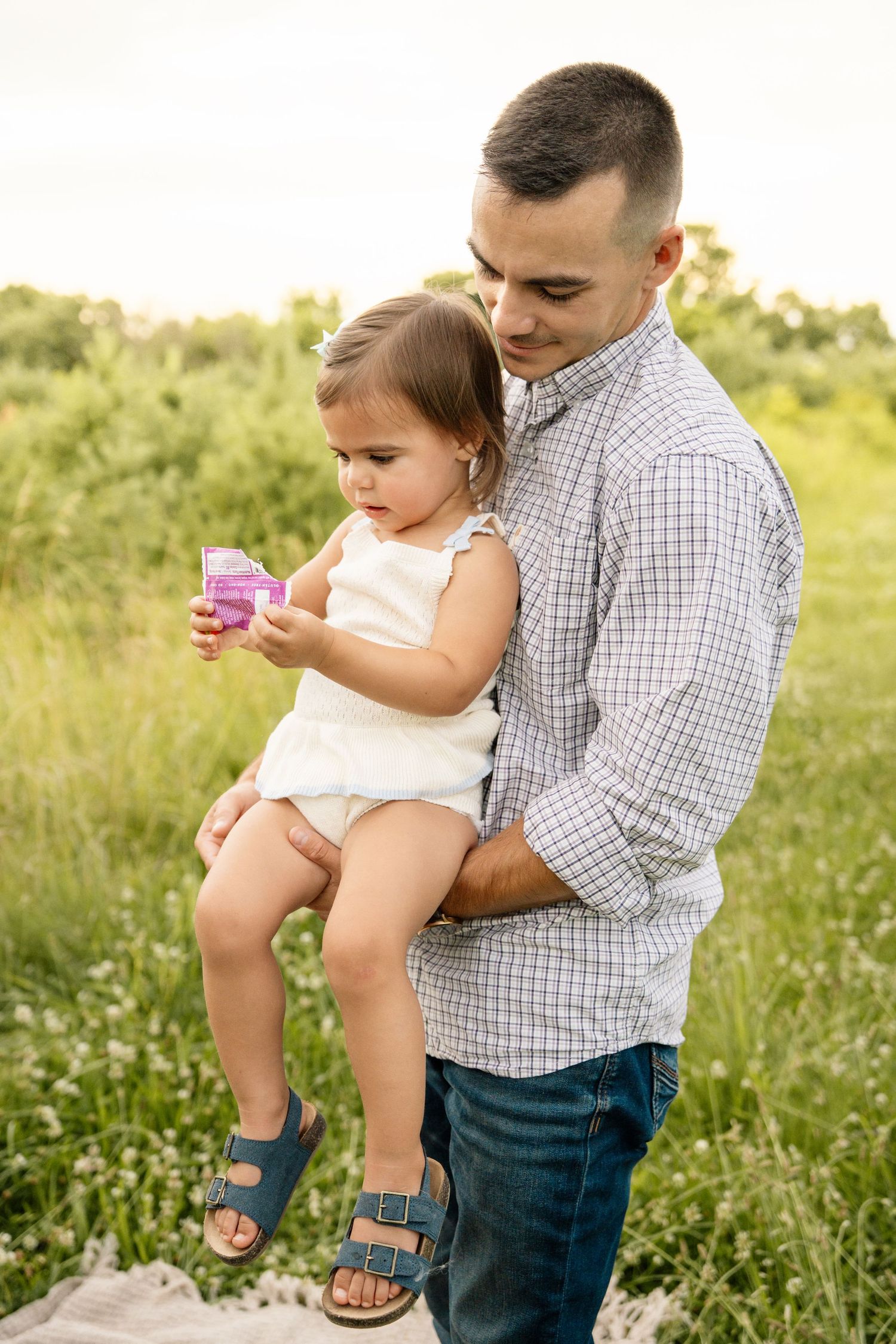Father holding his toddler daughter in a sunny green field as she plays with a small pink toy.