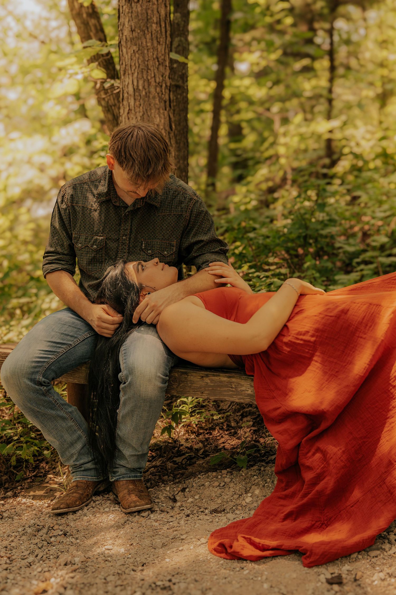 A pregnant woman in a red dress lies on a man's lap on a forest bench in a romantic maternity photo session.