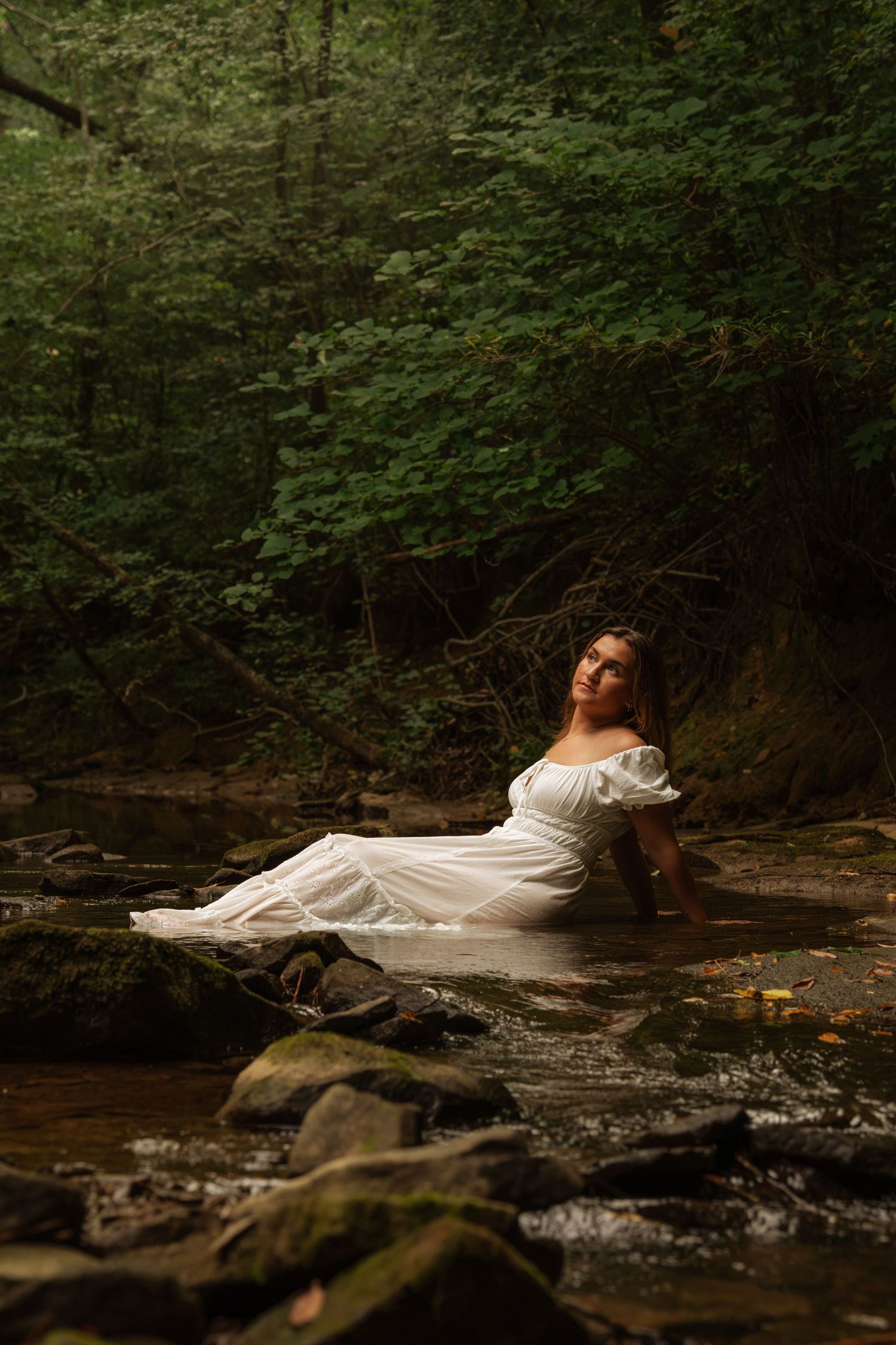 Woman in white dress sitting in a shallow rocky creek surrounded by green forest.