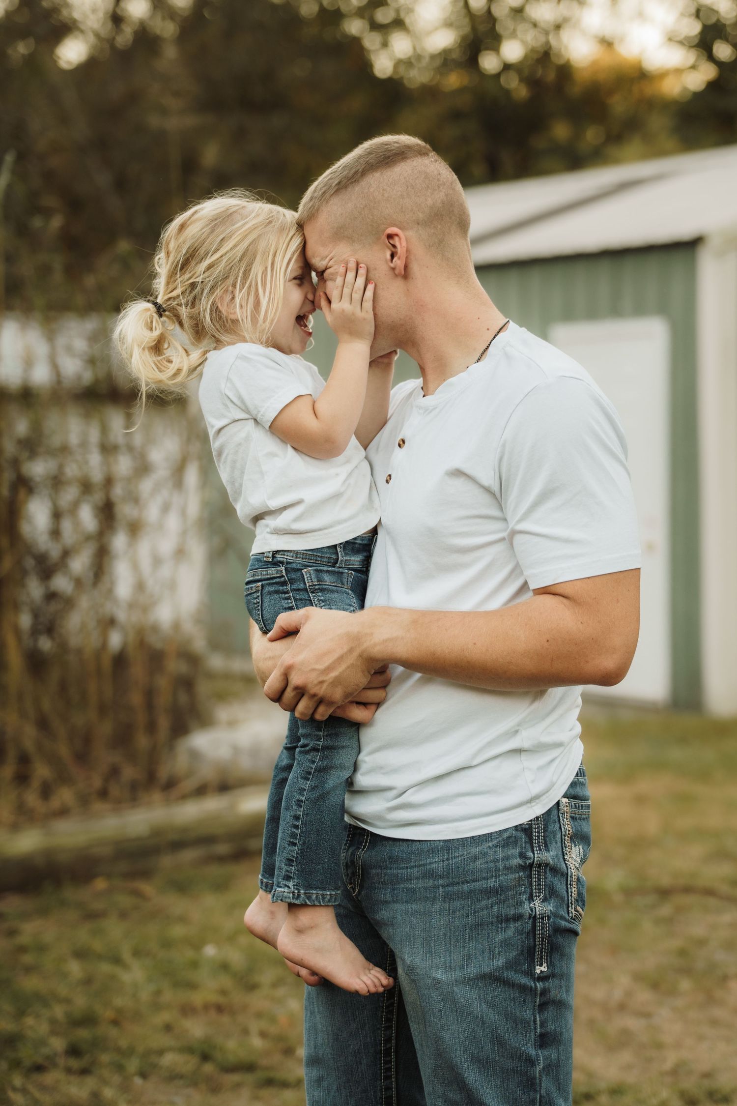 Father tenderly holds young blonde daughter as they touch foreheads and smile outdoors.