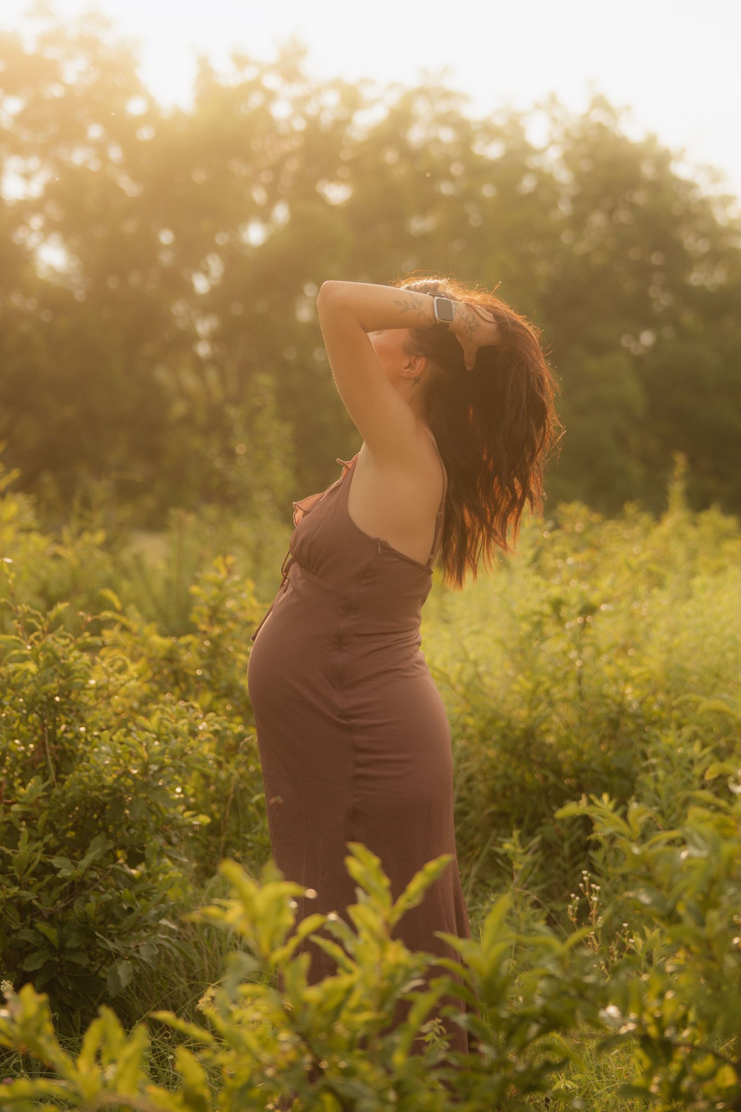 A woman in a brown dress stands among lush green plants, bathed in warm golden sunlight during a serene outdoor photoshoot.