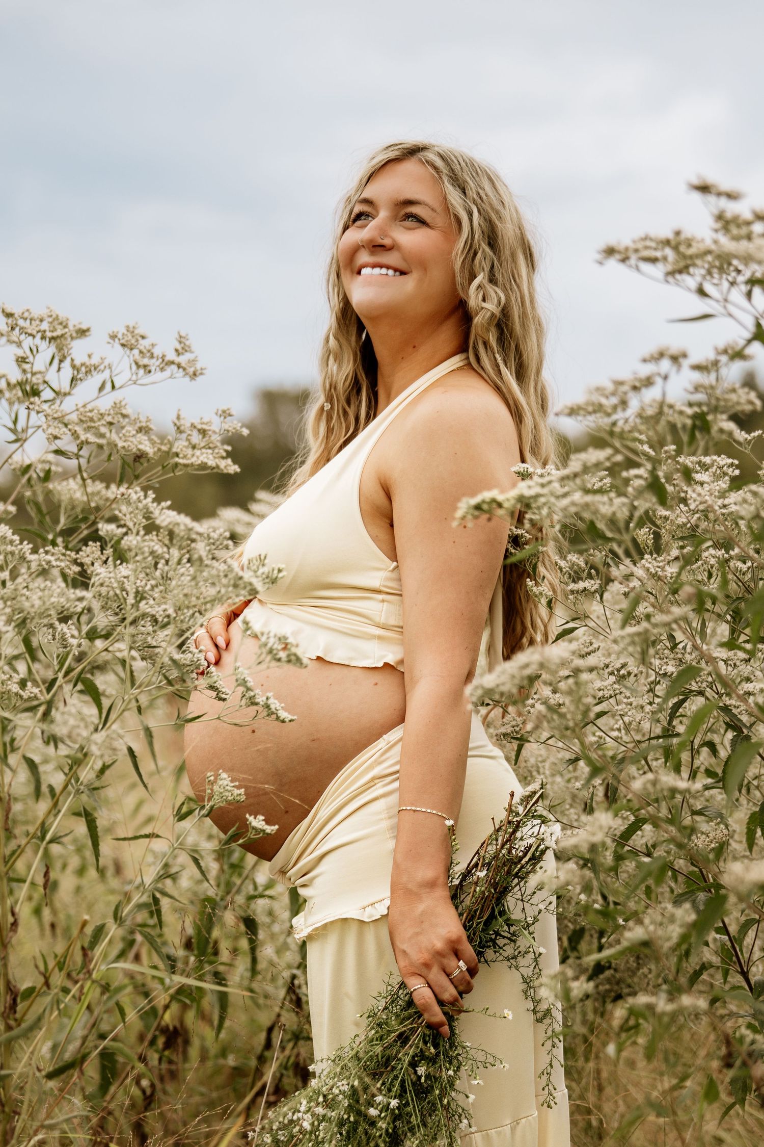 Smiling pregnant woman in yellow crop top holding wildflowers in blooming meadow.