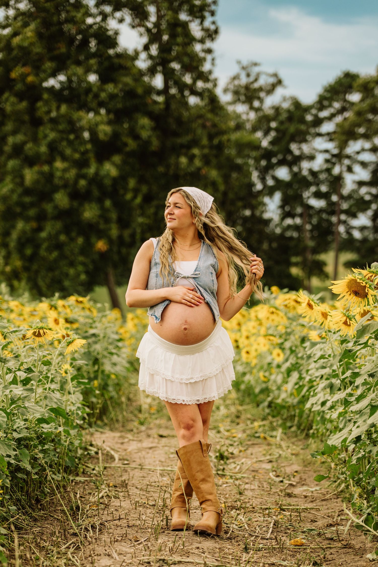 Pregnant woman in sunflower field wearing white skirt and cowboy boots, cradling baby bump outdoors.
