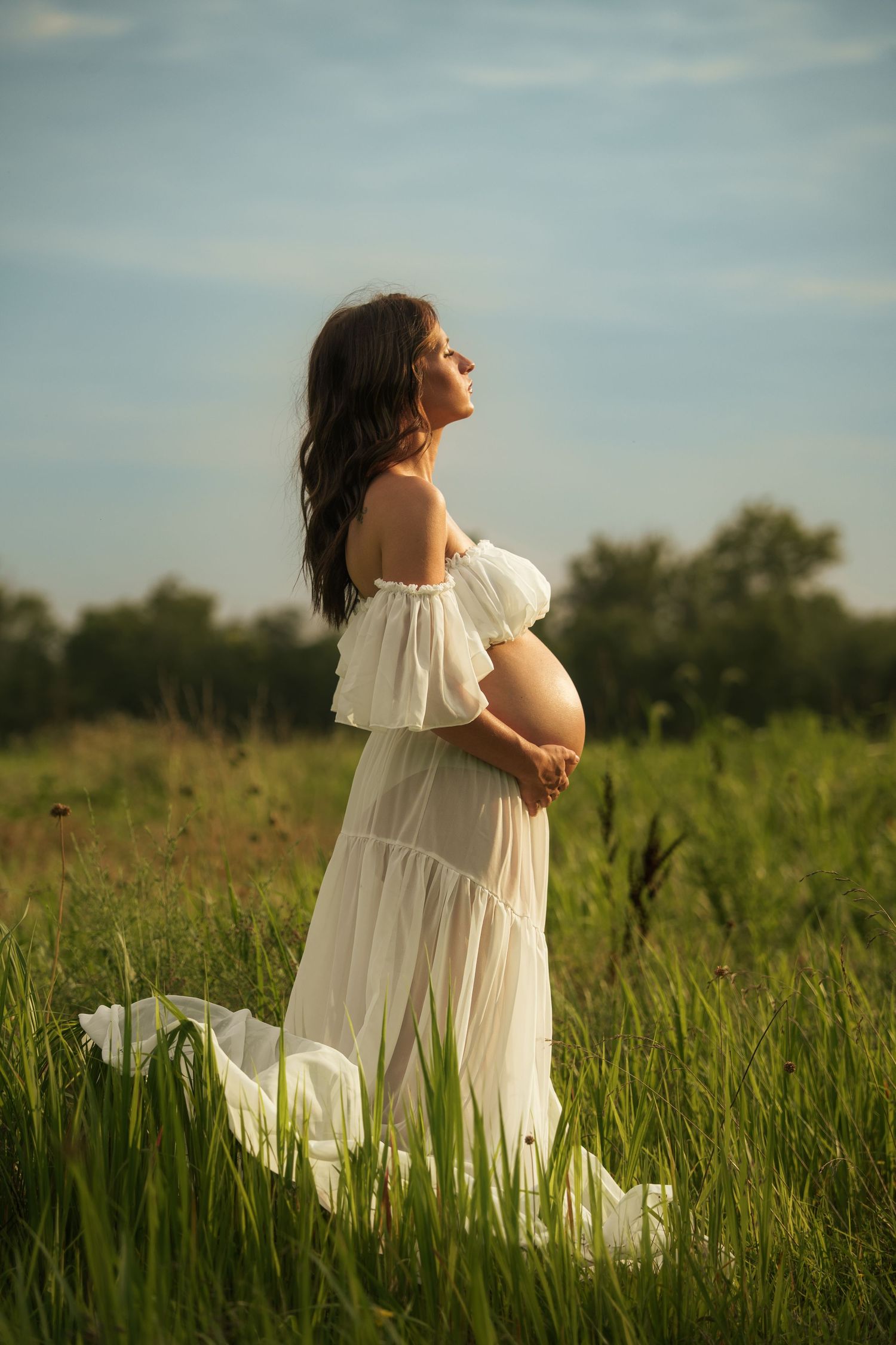 Side profile of pregnant woman in white dress gazing upward in a scenic outdoor field.