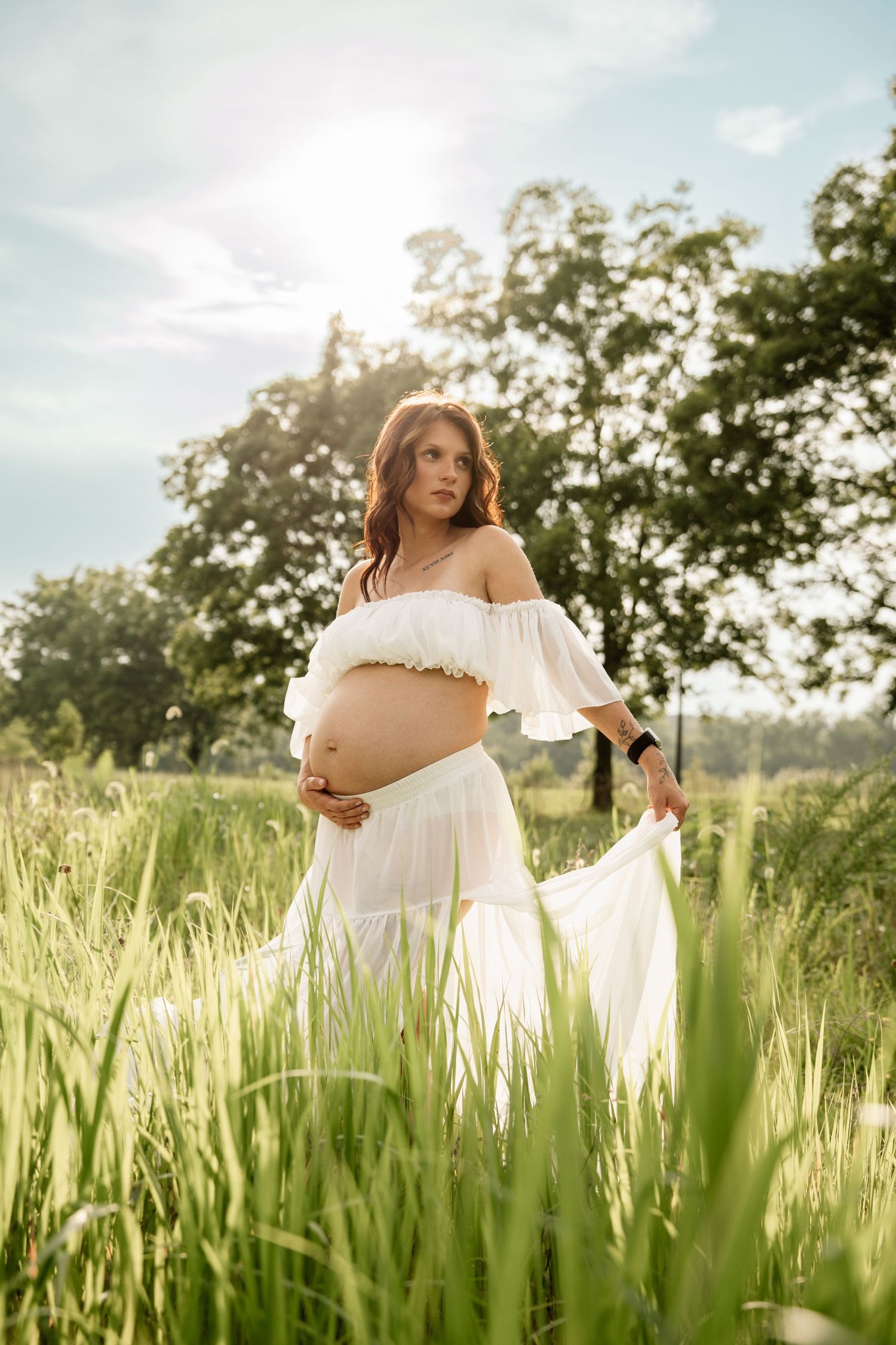 Pregnant woman in sheer white dress cradles baby bump while standing in tall grass with sunlight behind her.