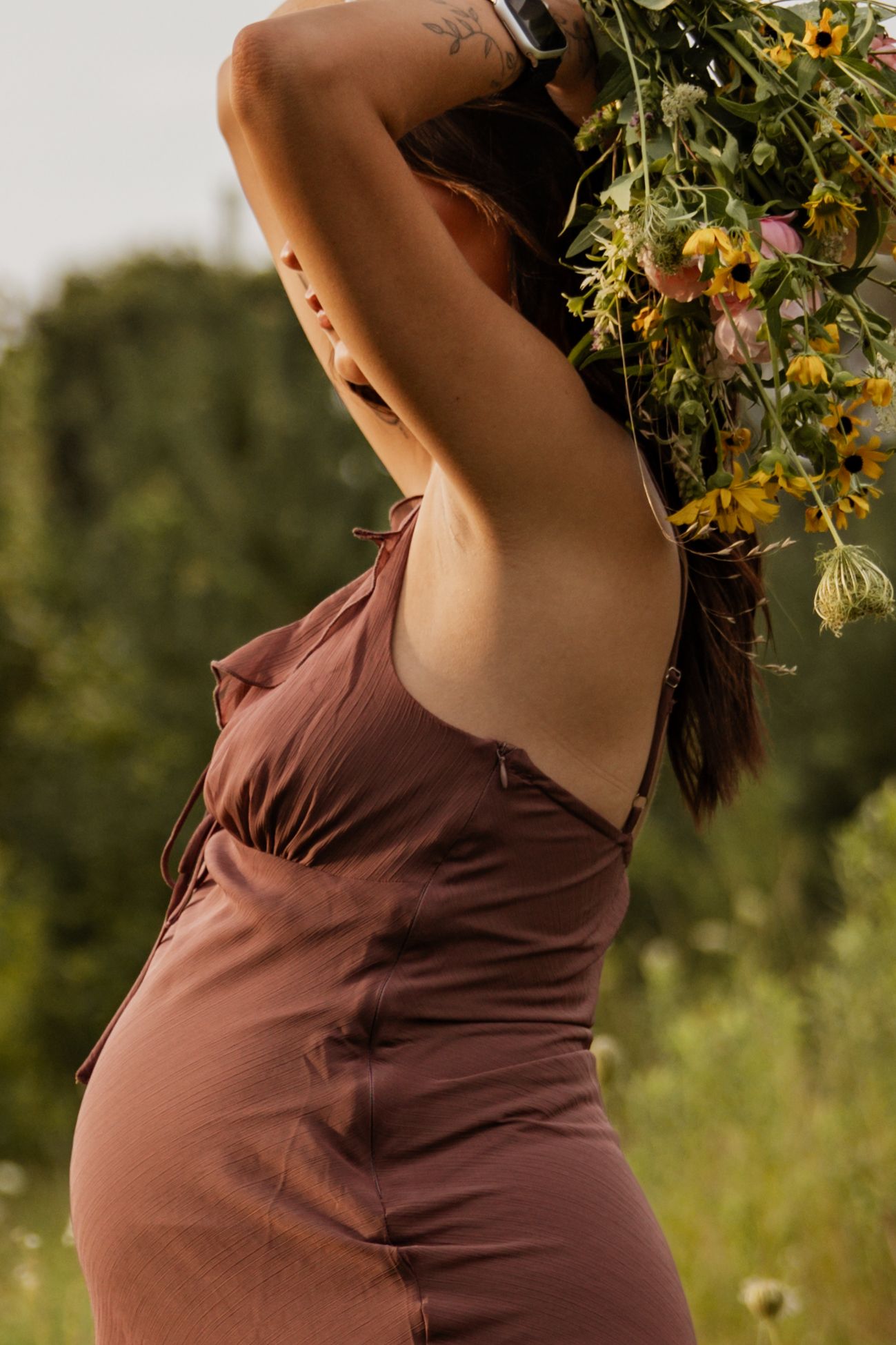 Pregnant woman in brown slip dress holds wildflower bouquet overhead showing back tattoo in sunny field.